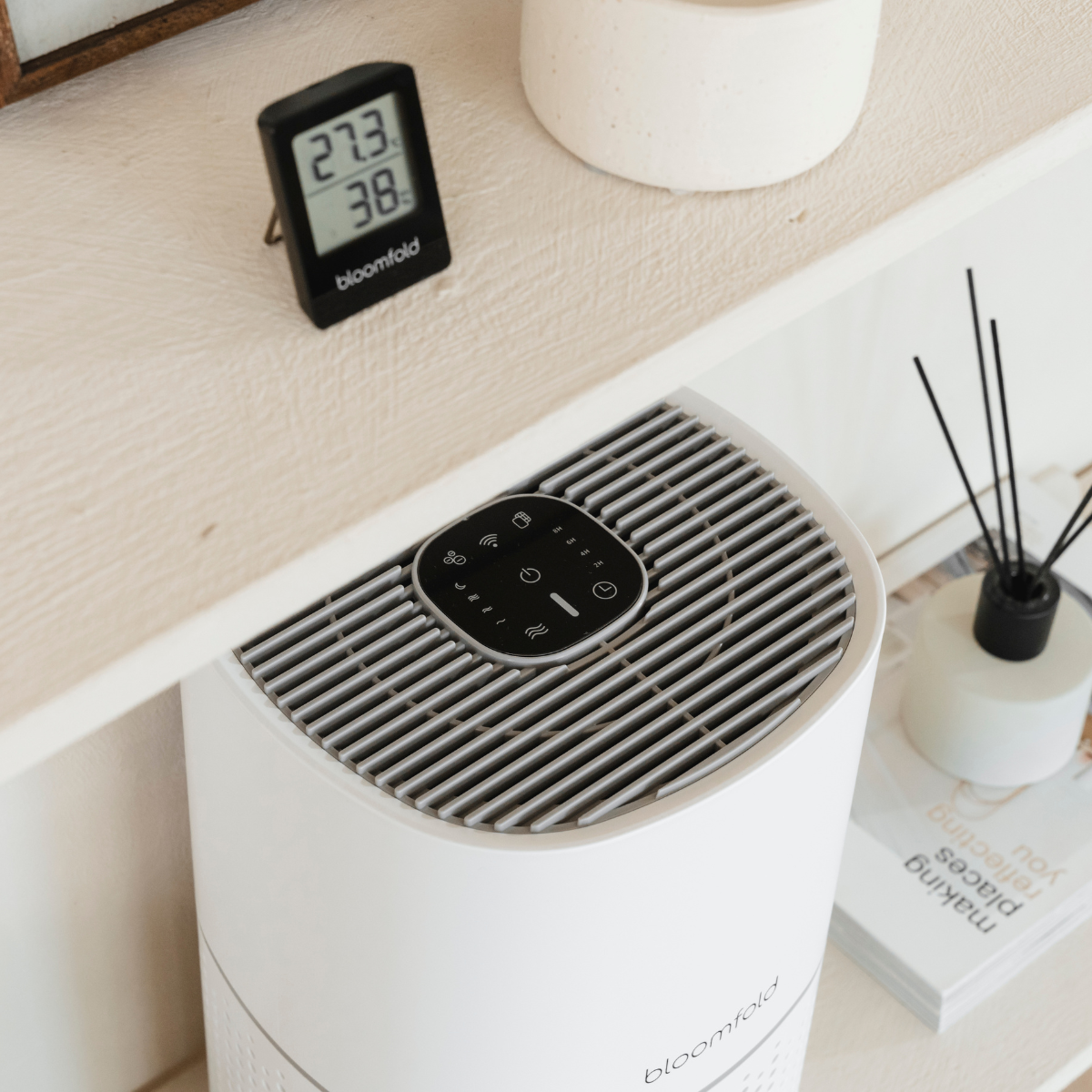 Purificatore d'aria bianco con display digitale in alto. C'è un termometro e un diffusore di canne nelle vicinanze.