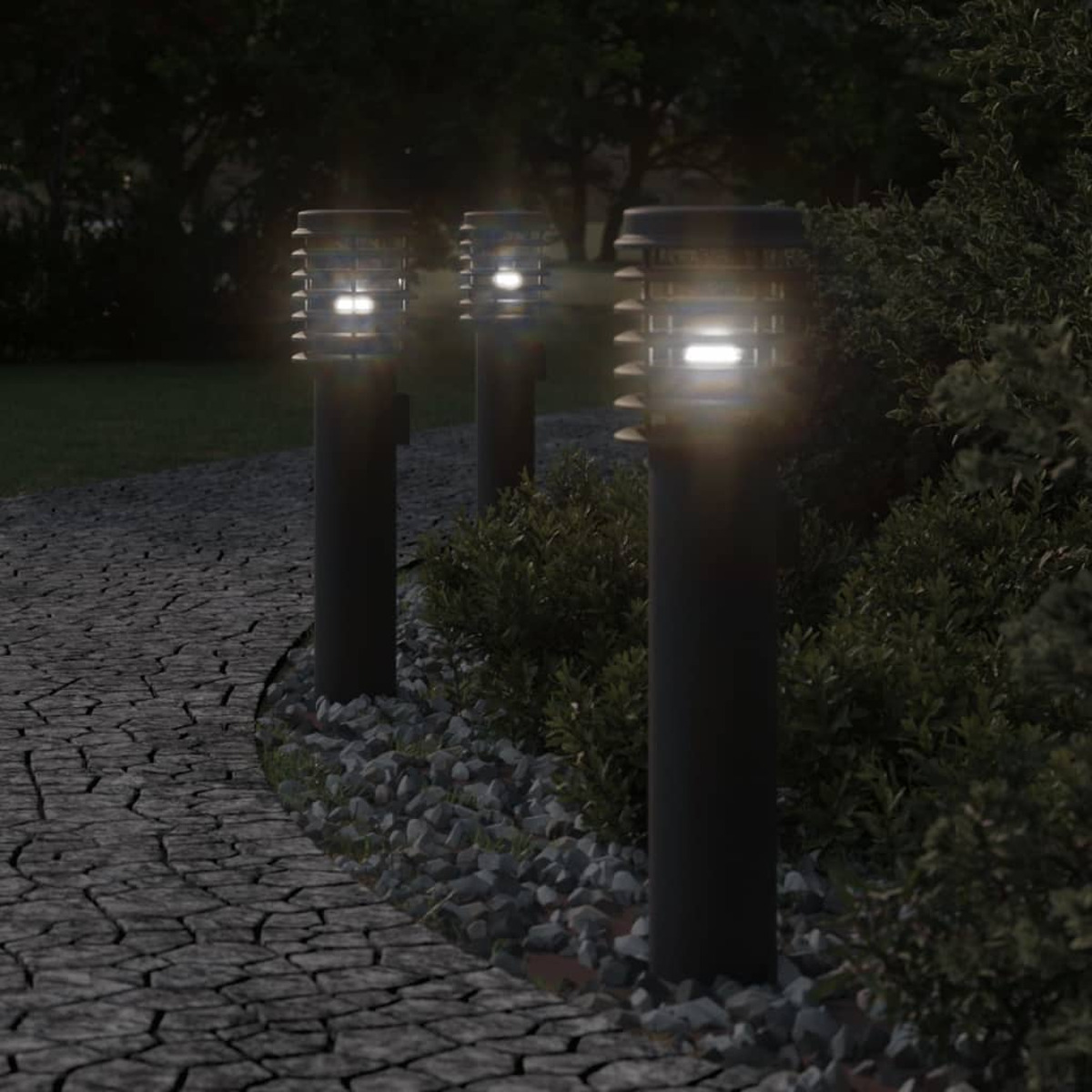 Drei dunkle, zylindrische Außenleuchten in einem Garten. Sie stehen auf einem Steinweg, mit Büschen.
