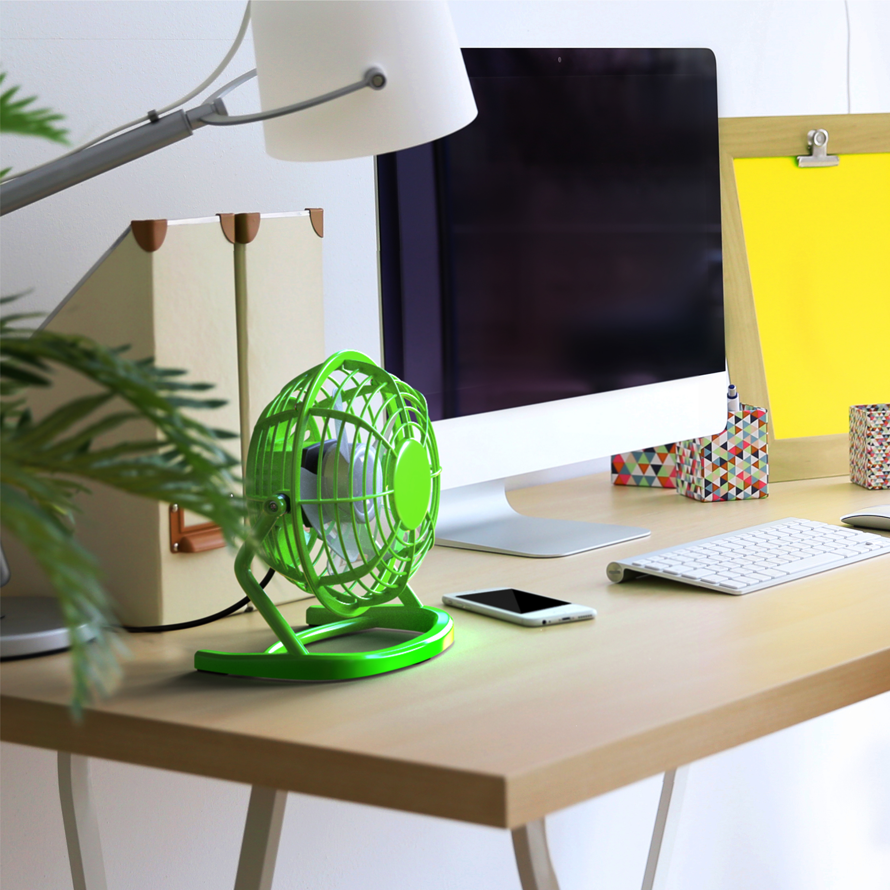 Grüner Ventilator auf einem Holztisch mit Computer, Lampe, Telefon und Tastatur. Grüne Pflanze daneben.