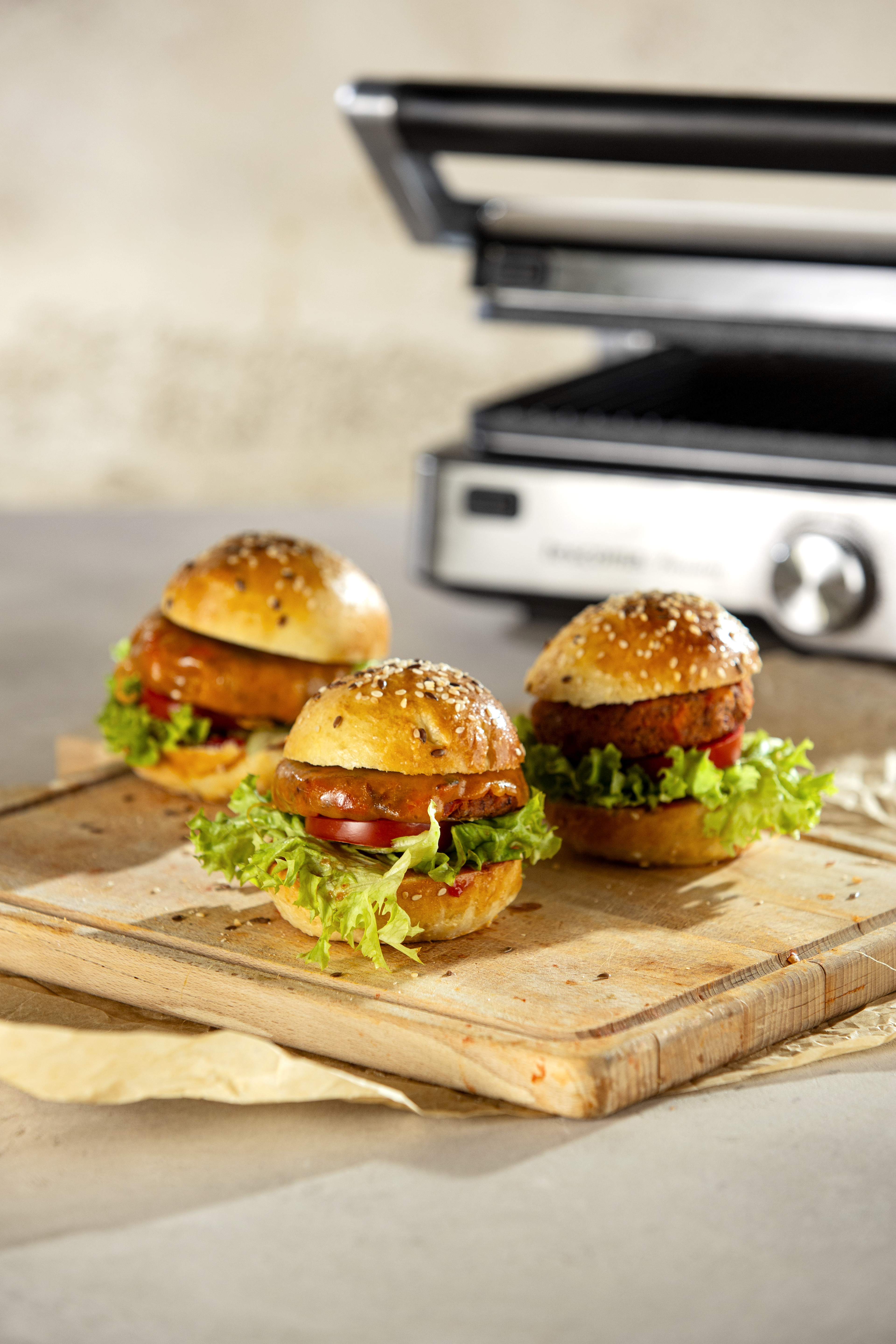 Drei Burger auf einem Holzbrett mit Grill im Hintergrund. Sie haben Sesambrötchen und Salat.