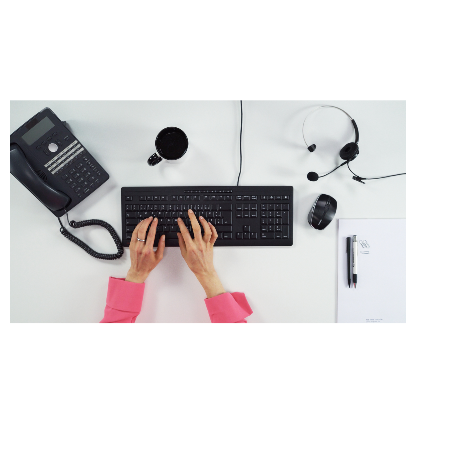 Widok z góry na białe biurko. Zawiera telefon, klawiaturę, zestaw słuchawkowy i papier.