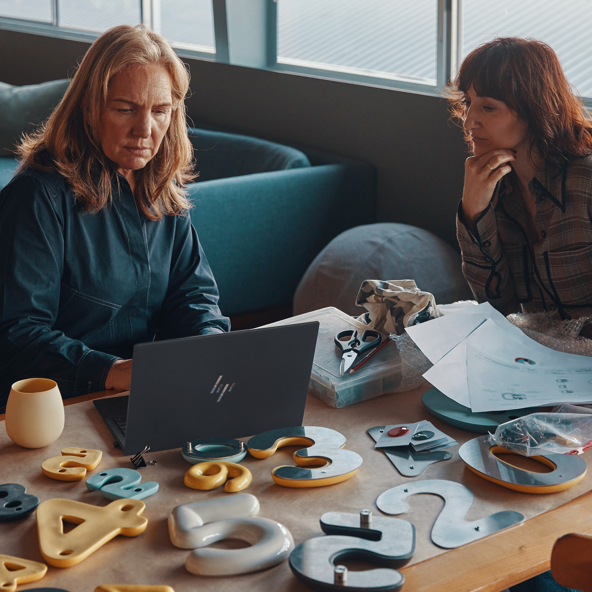 Due donne a tavola con laptop. Carte e strumenti. Una guarda il laptop, l'altra le carte.