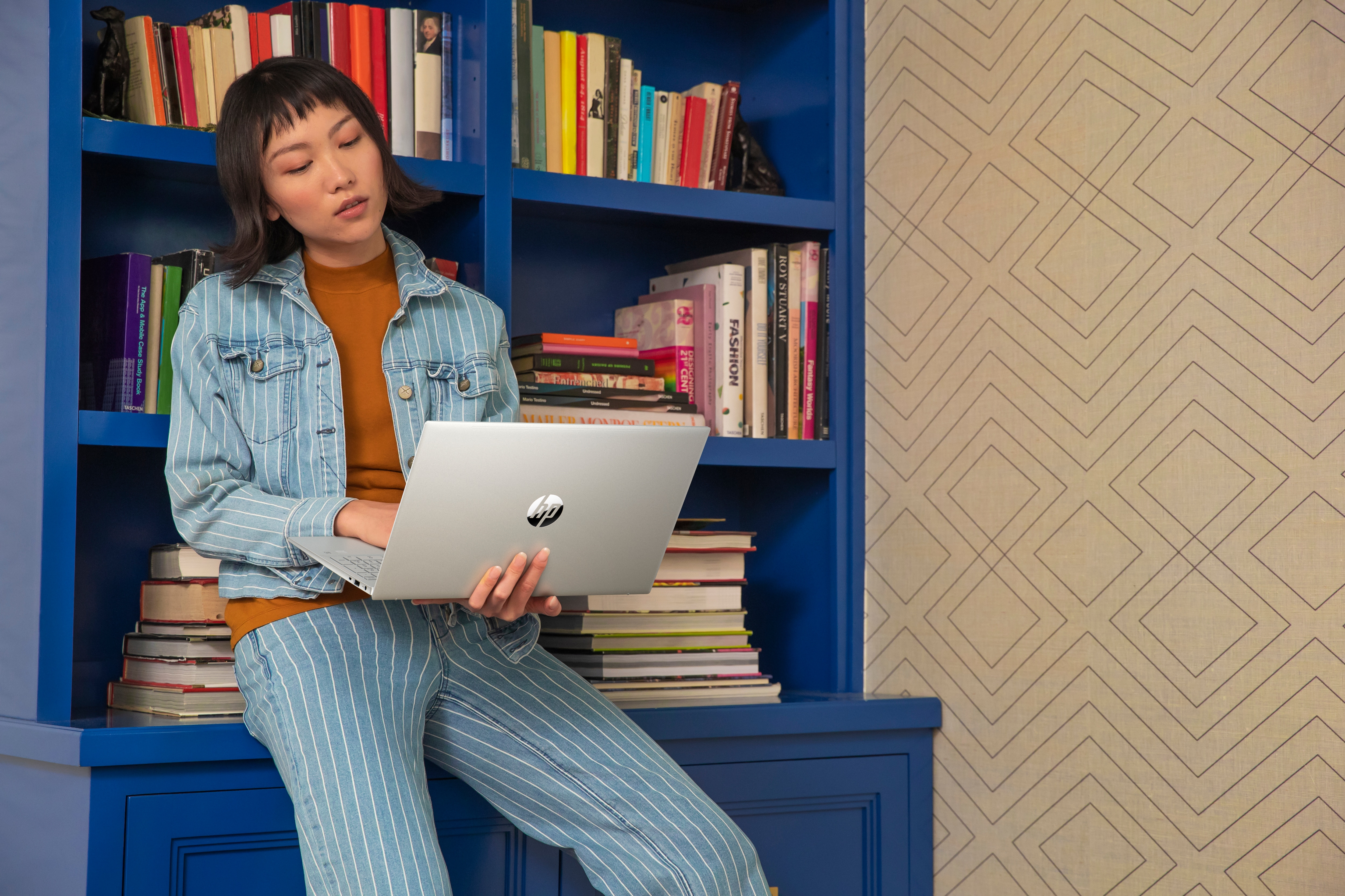 Una donna usa un laptop davanti a una libreria blu. Indossa una giacca di jeans e pantaloni a righe.