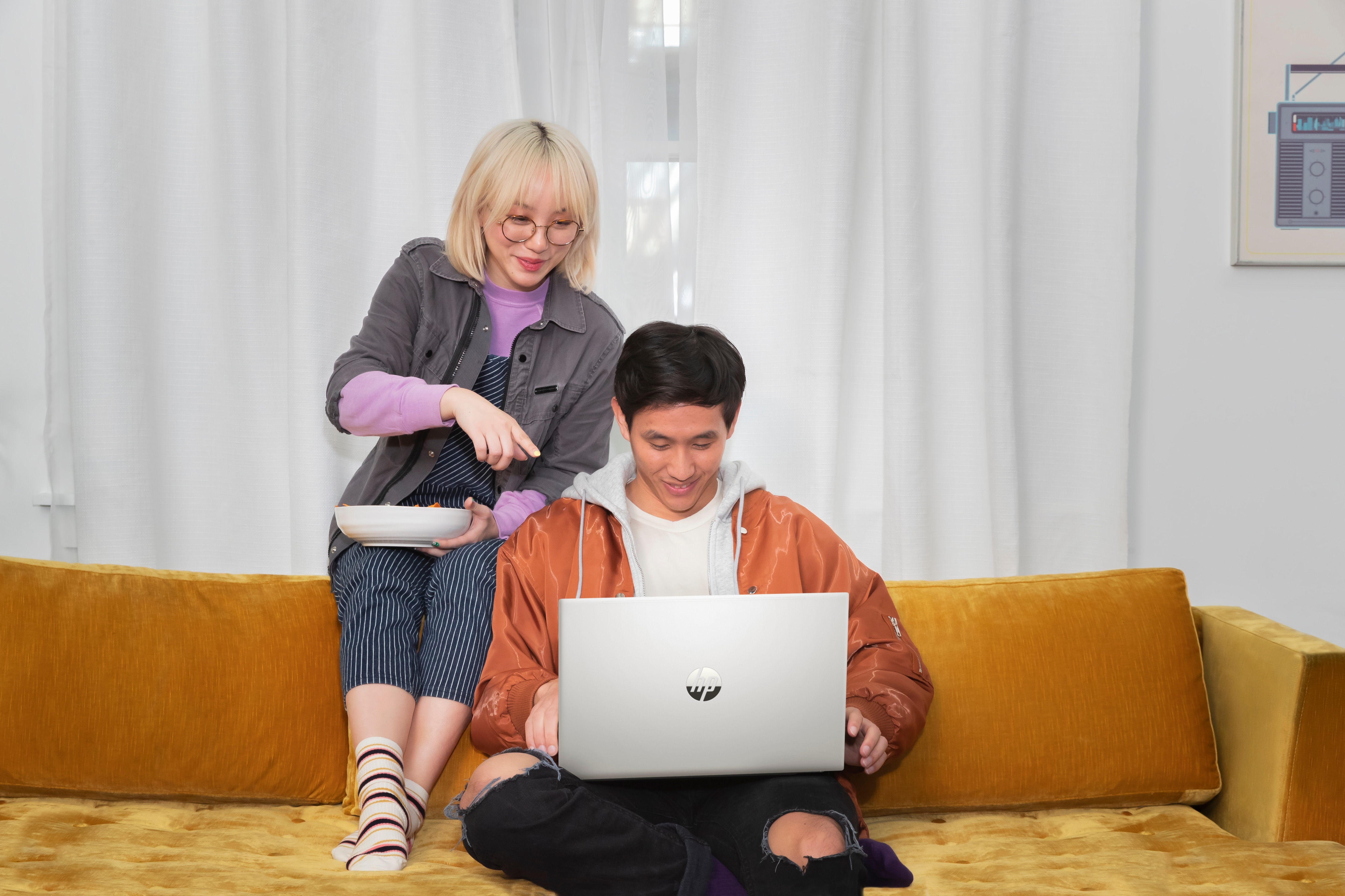 Dwie osoby na żółtej kanapie korzystające z laptopa. Kobieta wskazuje na ekran.