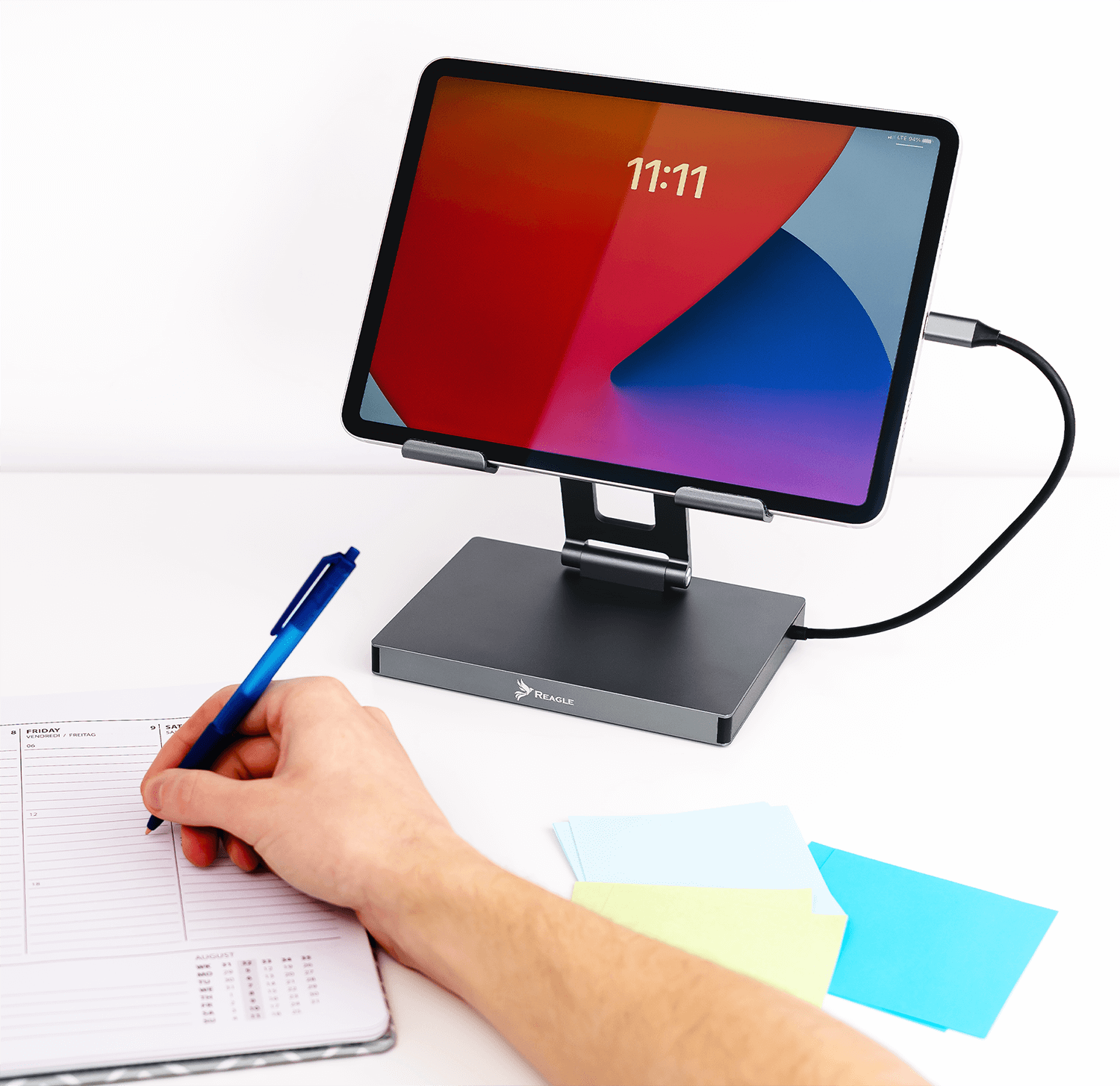 Ręka pisząca w planerze obok tabletu na stojaku z kablem.