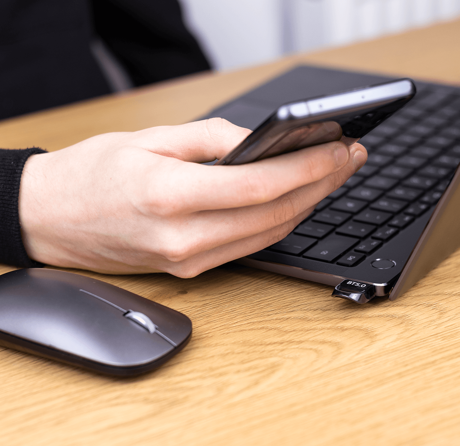 Dłonie osoby trzymającej telefon nad klawiaturą, z myszą na drewnianym biurku.