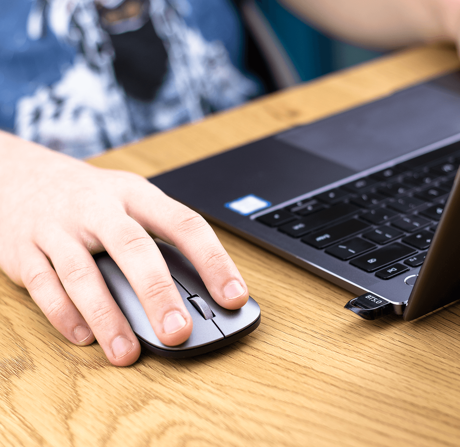 Dłoń używająca szarej myszy na drewnianym biurku obok laptopa.