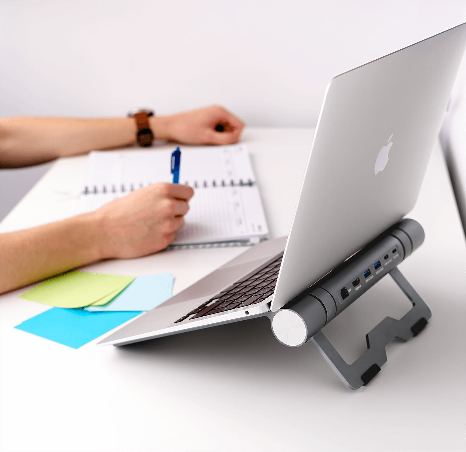Laptop na stojaku, z długopisem, notatnikiem i karteczkami na biurku. Ręka pisze.