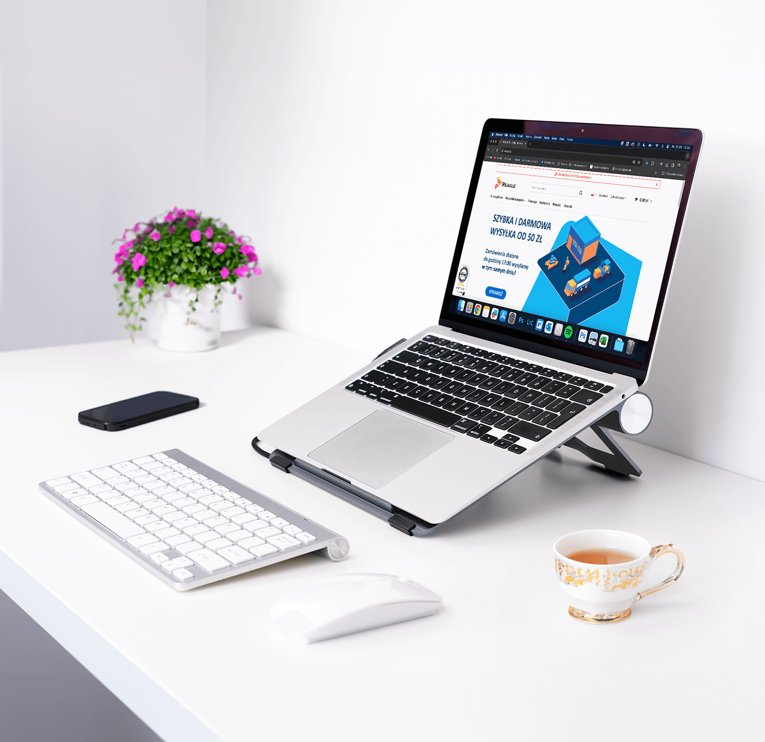 Białe biurko z laptopem na stojaku, klawiaturą, telefonem, myszką i filiżanką z kwiatami.