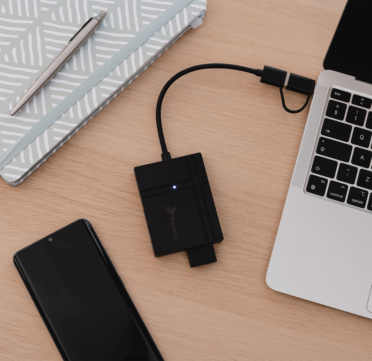 Laptop, telefon i czarny czytnik kart SD połączone kablem, na drewnianej powierzchni.