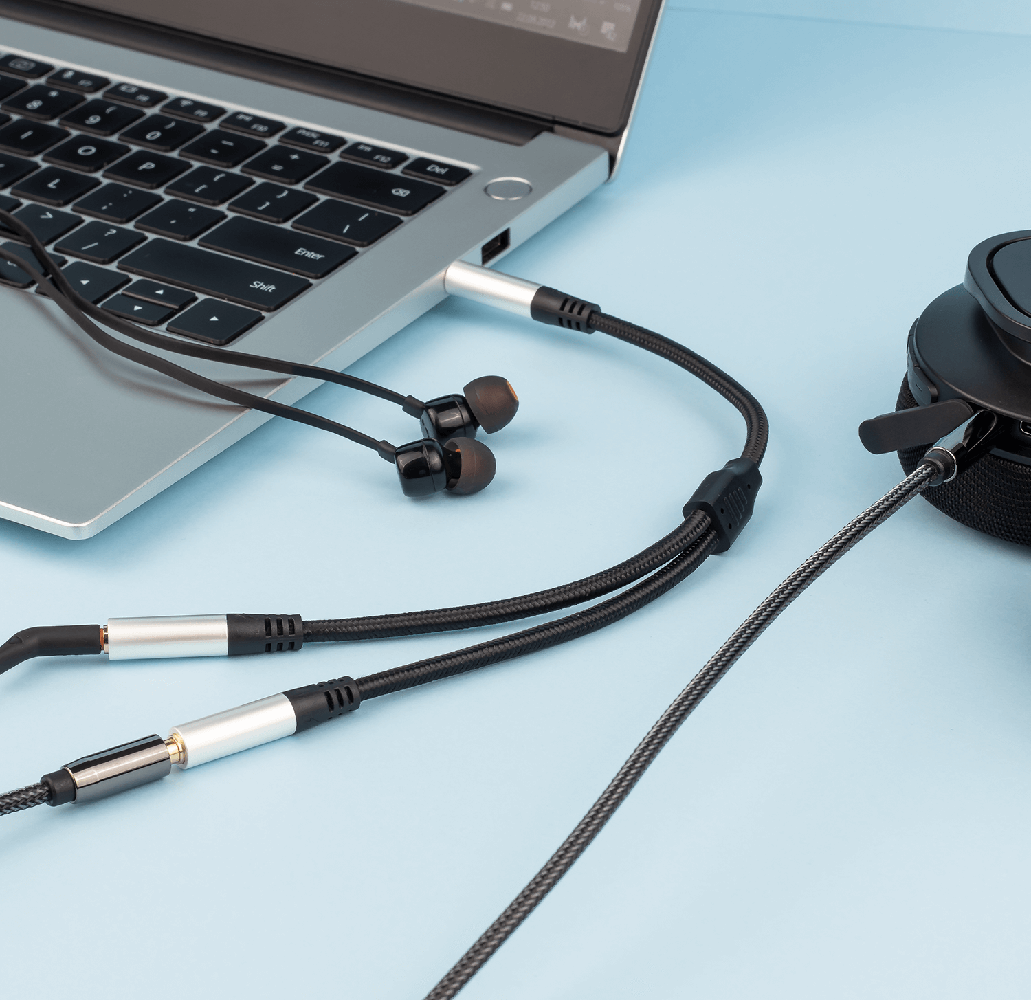 Laptop ze słuchawkami podłączonymi za pomocą rozdzielonego kabla, na jasnoniebieskiej powierzchni.