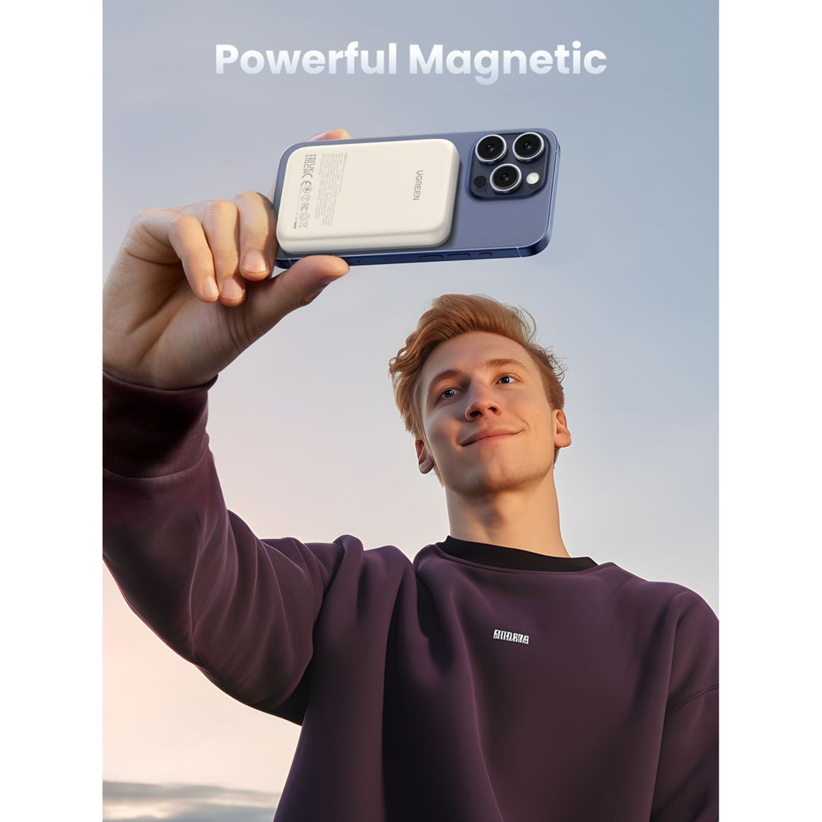 Mężczyzna trzymający telefon z dołączonym magnetycznym power bankiem. Widoczne błękitne niebo i marka Ugreen.