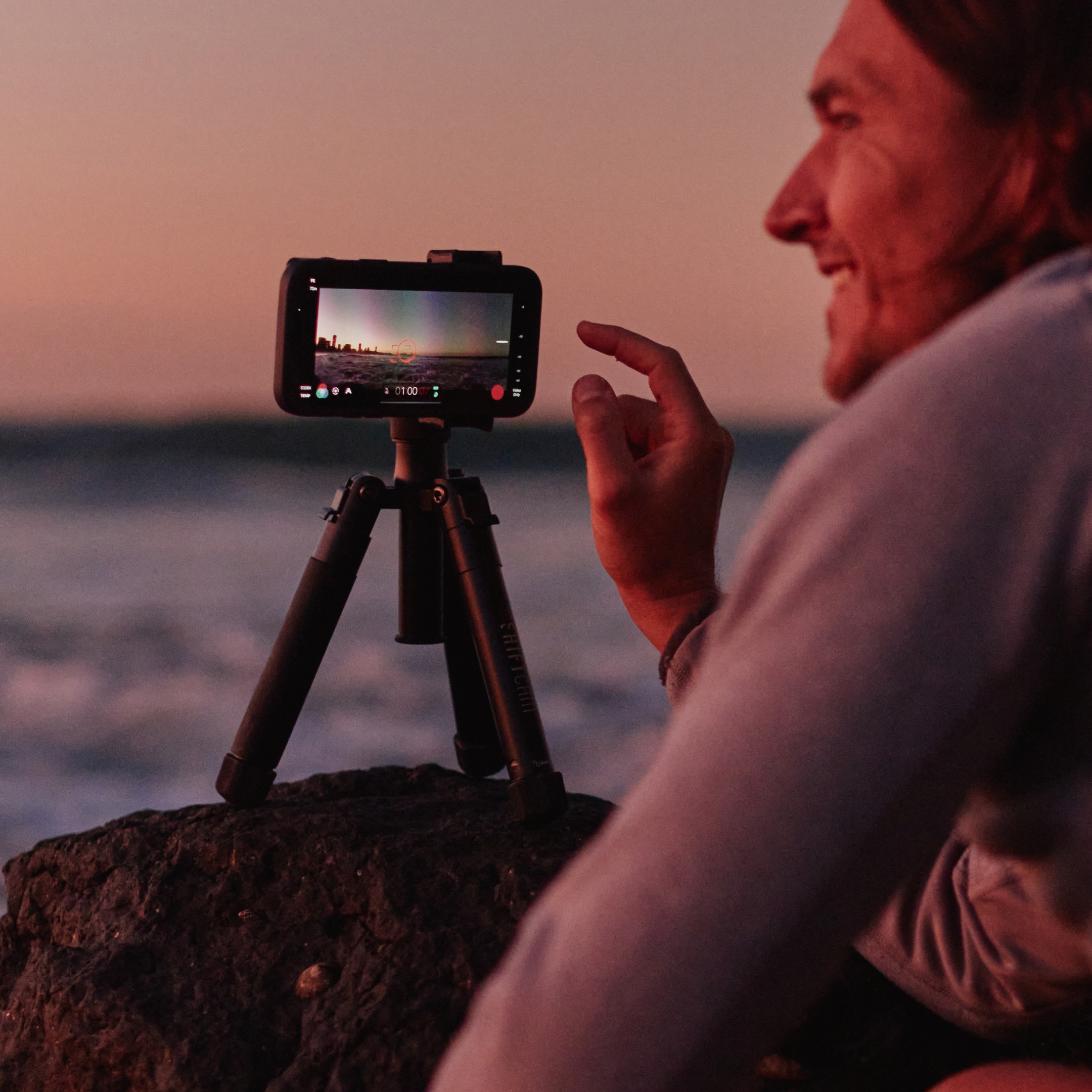 Mann benutzt Telefonkamera auf Stativ am Strand. Sonnenuntergang im Hintergrund.