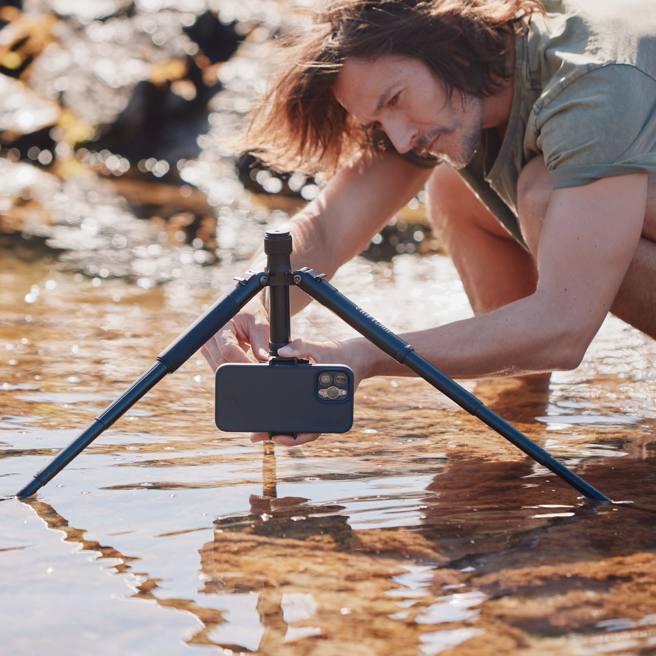 Mann richtet ein Telefon auf einem Stativ in flachem Wasser ein, mit unscharfem Hintergrund.