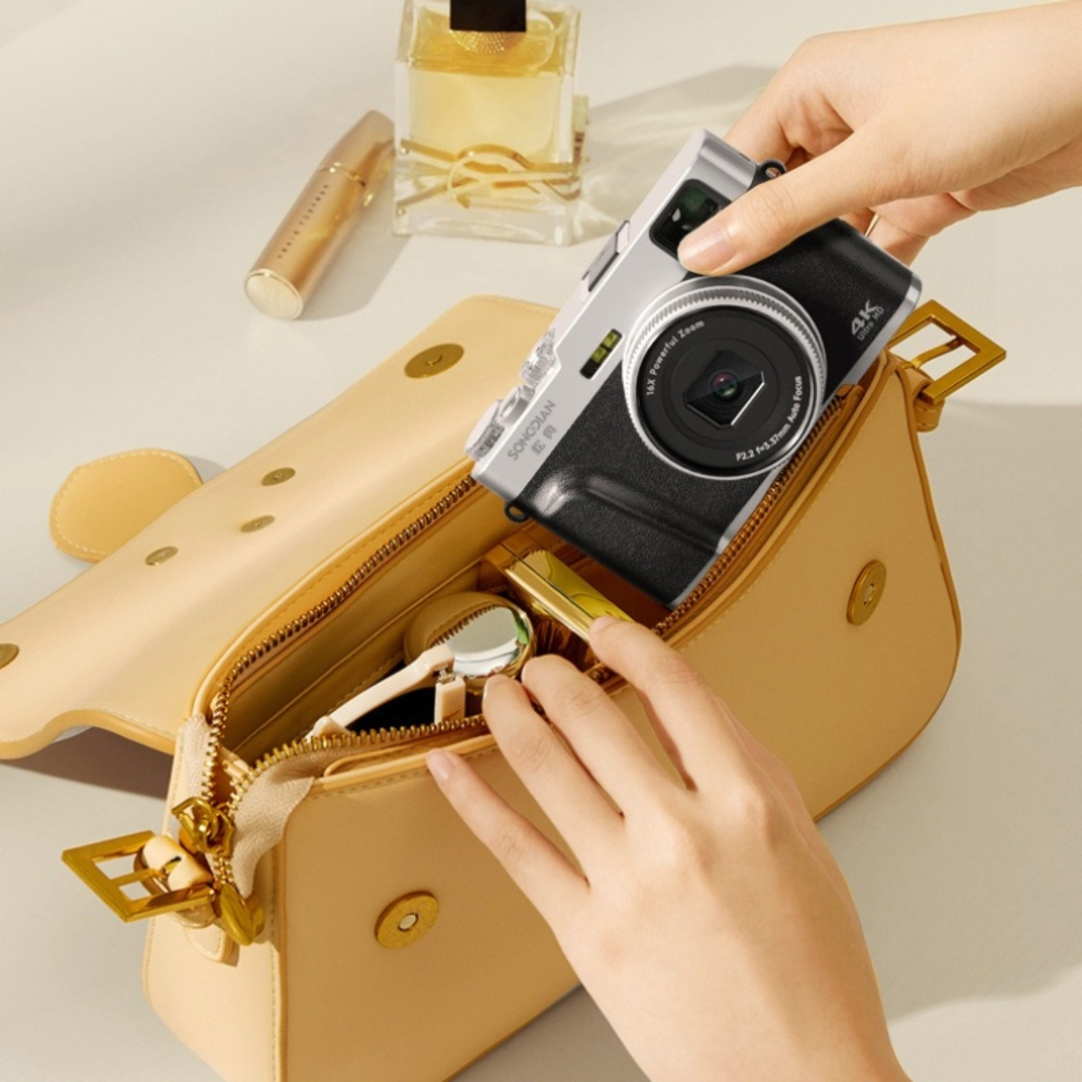 Hände legen eine Kamera in eine beige Tasche. Lippenstift, Parfüm und Spiegel sind sichtbar.