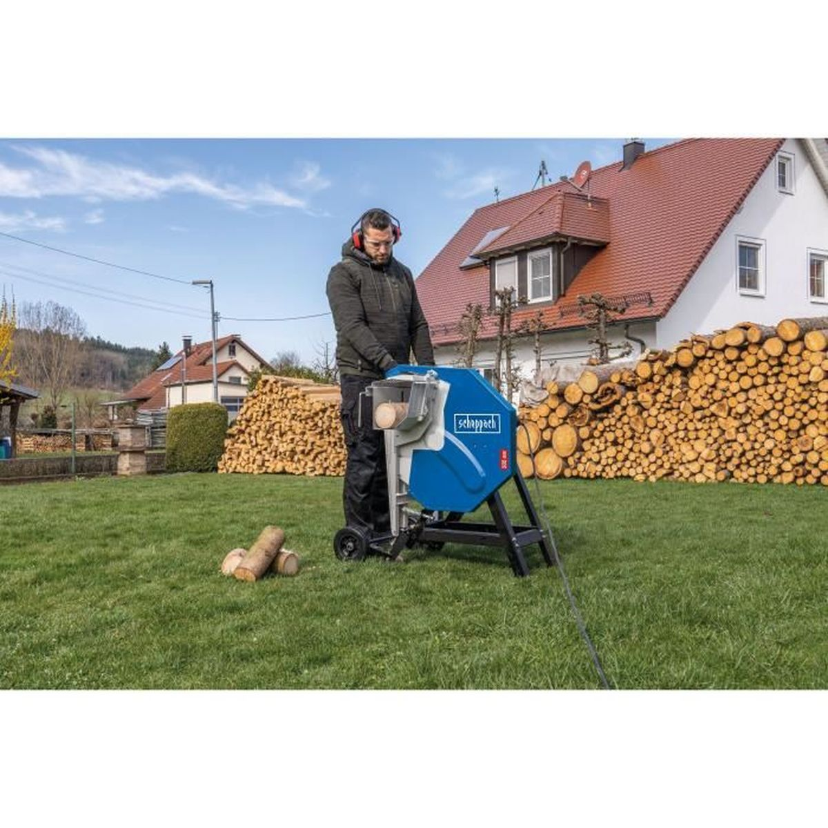 Ein Mann benutzt eine blaue Holzsäge auf einer Wiese, im Hintergrund gestapelte Baumstämme.