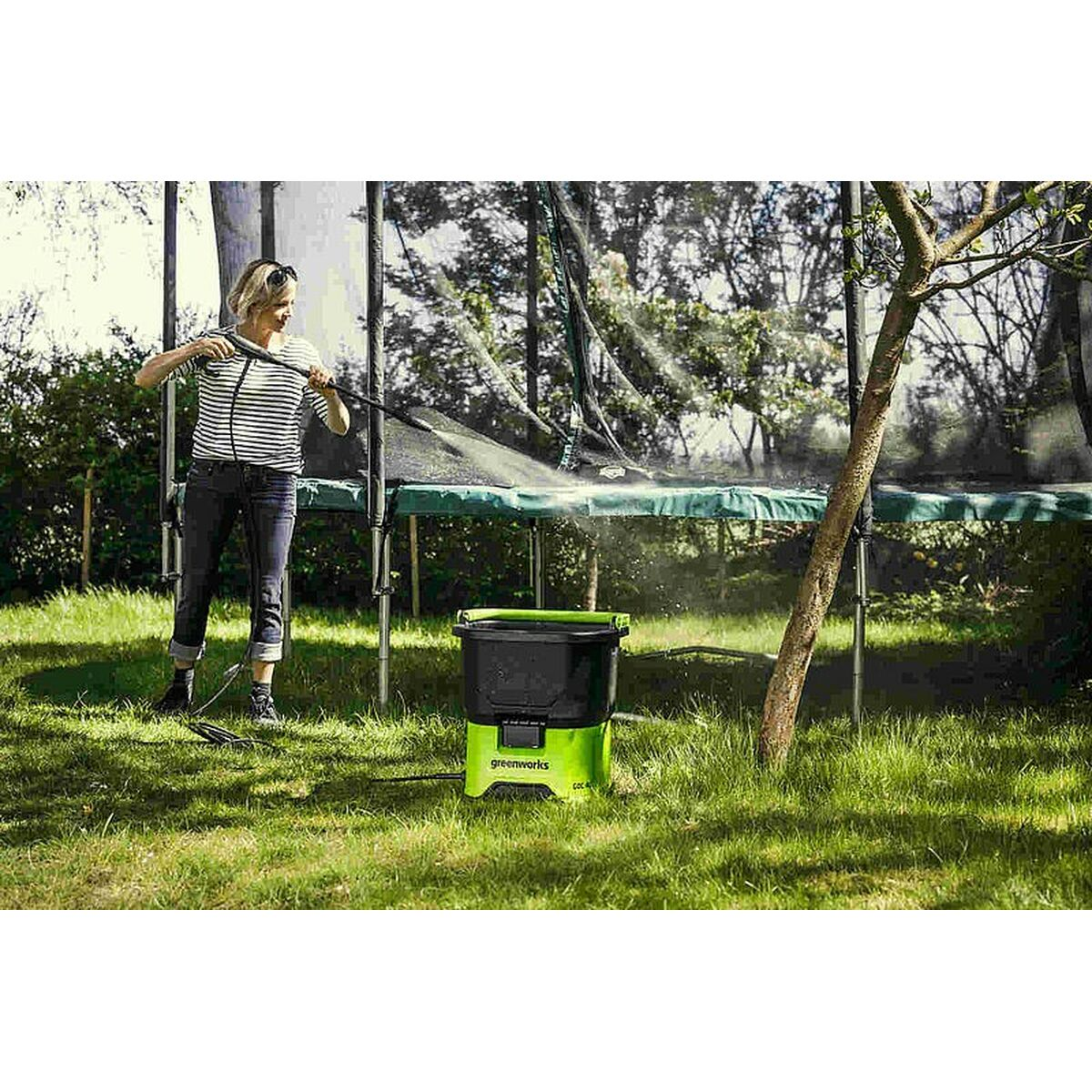 Eine Frau reinigt ein Trampolin mit einem Hochdruckreiniger auf einer Wiese. Eine grüne Maschine steht daneben.