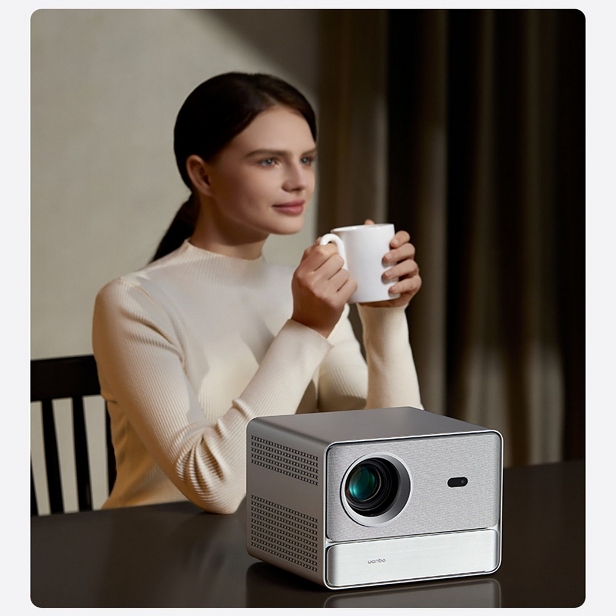 Mujer sosteniendo una taza, proyector en la mesa. La mujer está en una habitación con luz tenue.