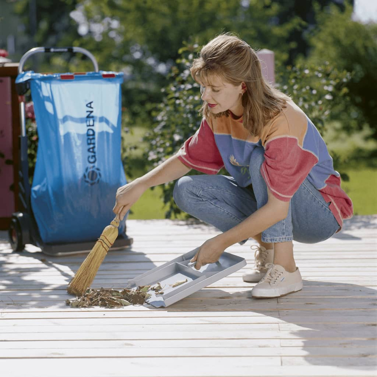 Frau kehrt eine Holzterrasse. Sie trägt Freizeitkleidung und benutzt einen Besen und eine Kehrschaufel auf dem Deck.