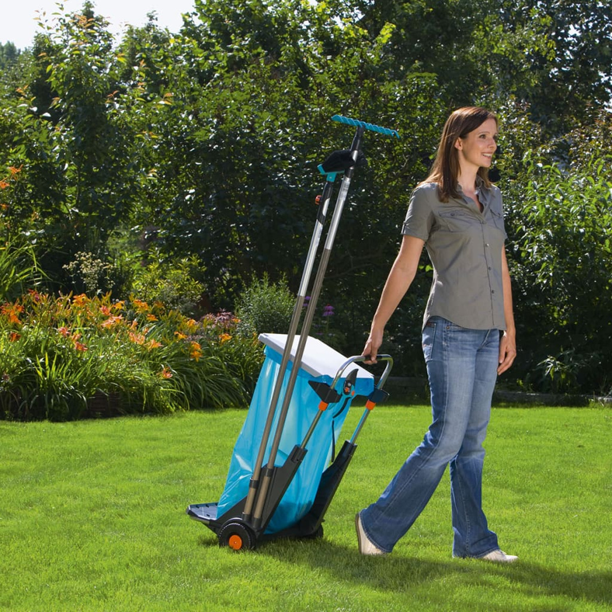 Frau geht mit einem Gartenwagen auf grünem Gras. Der Wagen ist blau mit schwarzen Rädern.