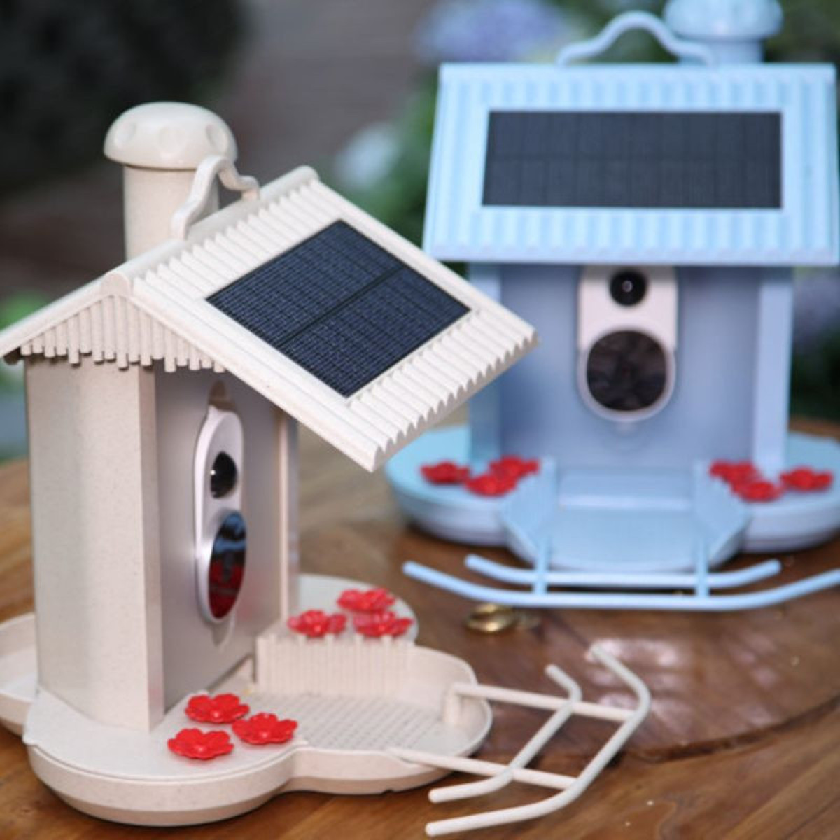 Zwei Vogelfutterhäuschen, Solarpanel und rote Blumen. Eines ist beige, eines hellblau. Draußen, sonnig.
