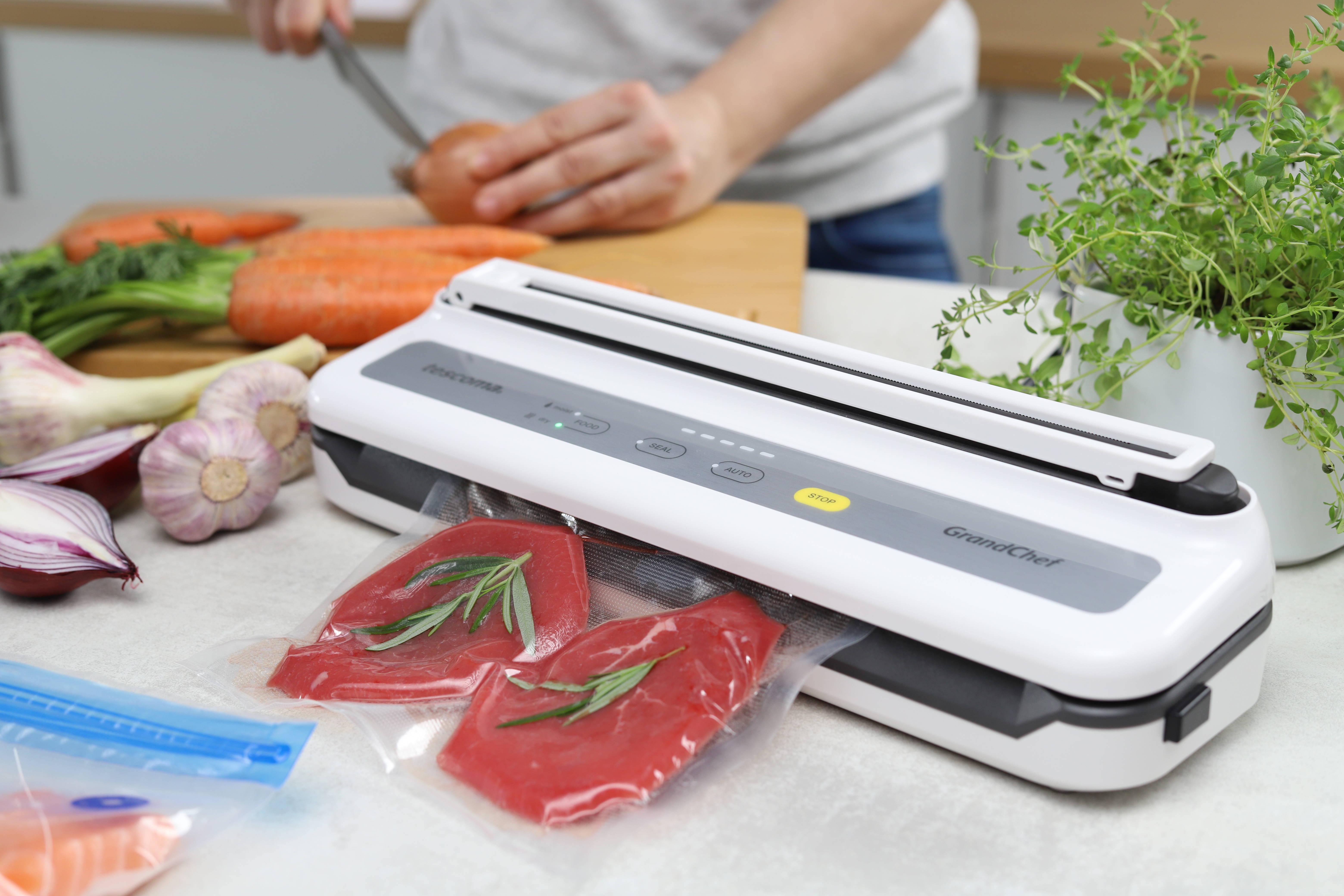 Ein Vakuumiergerät mit rohem Fleisch und Rosmarin, Gemüse auf einem Tisch und eine Person schneidet.