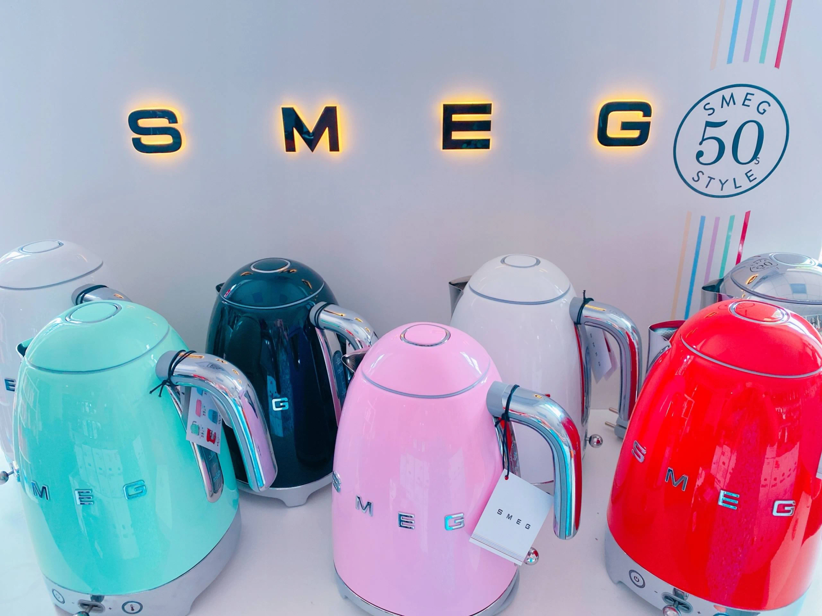 Kolekcja czajników SMEG w różnych kolorach z podświetlanym znakiem SMEG powyżej.