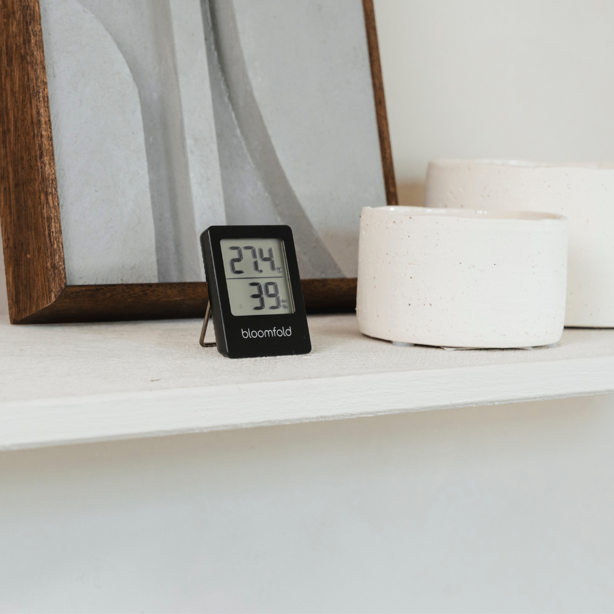 Ein schwarzes digitales Thermometer zeigt 27,4 und 39, platziert auf einem Regal. Daneben zwei weiße Keramiktöpfe.