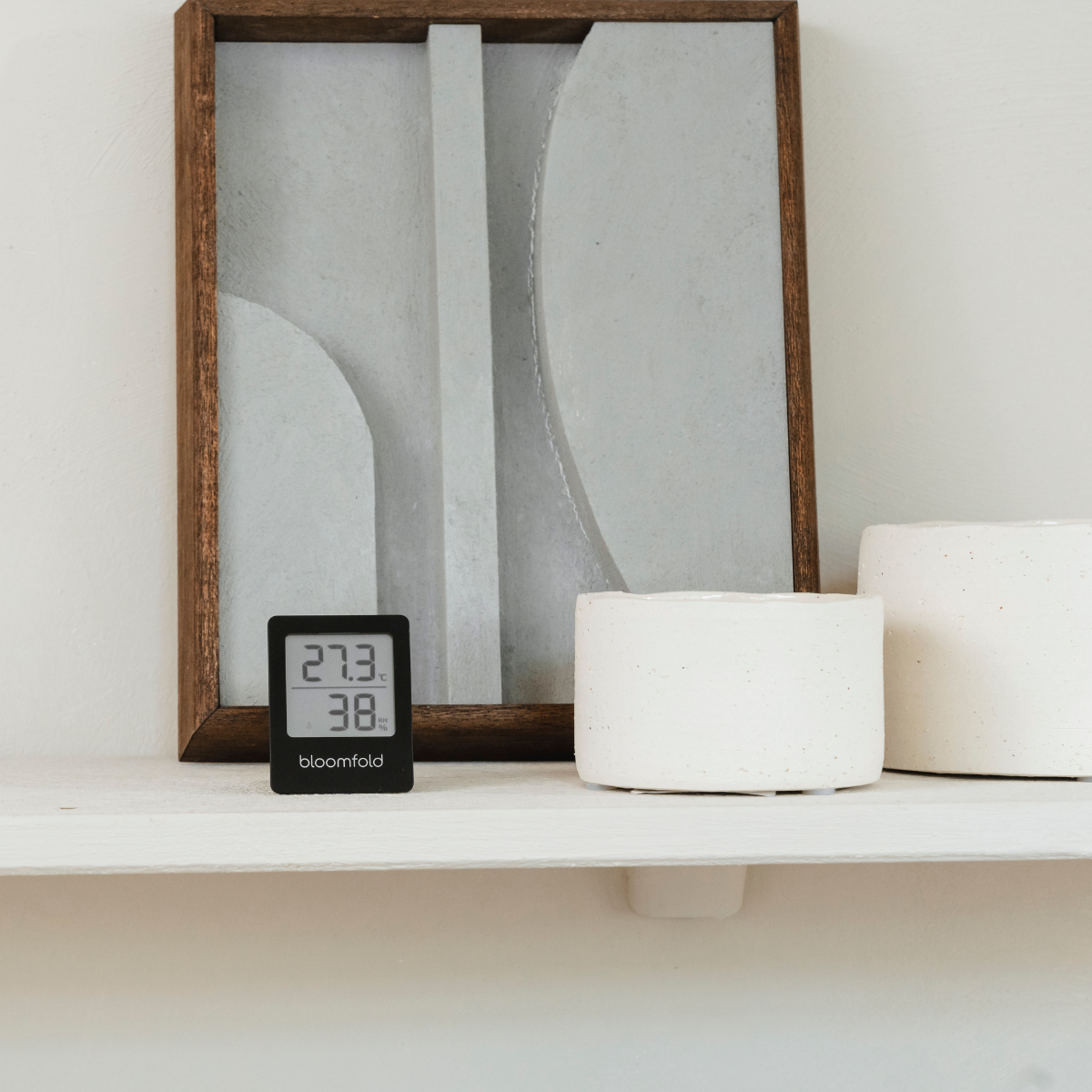 Kleines digitales Thermometer auf weißem Regal mit Kunst und weißen Töpfen.