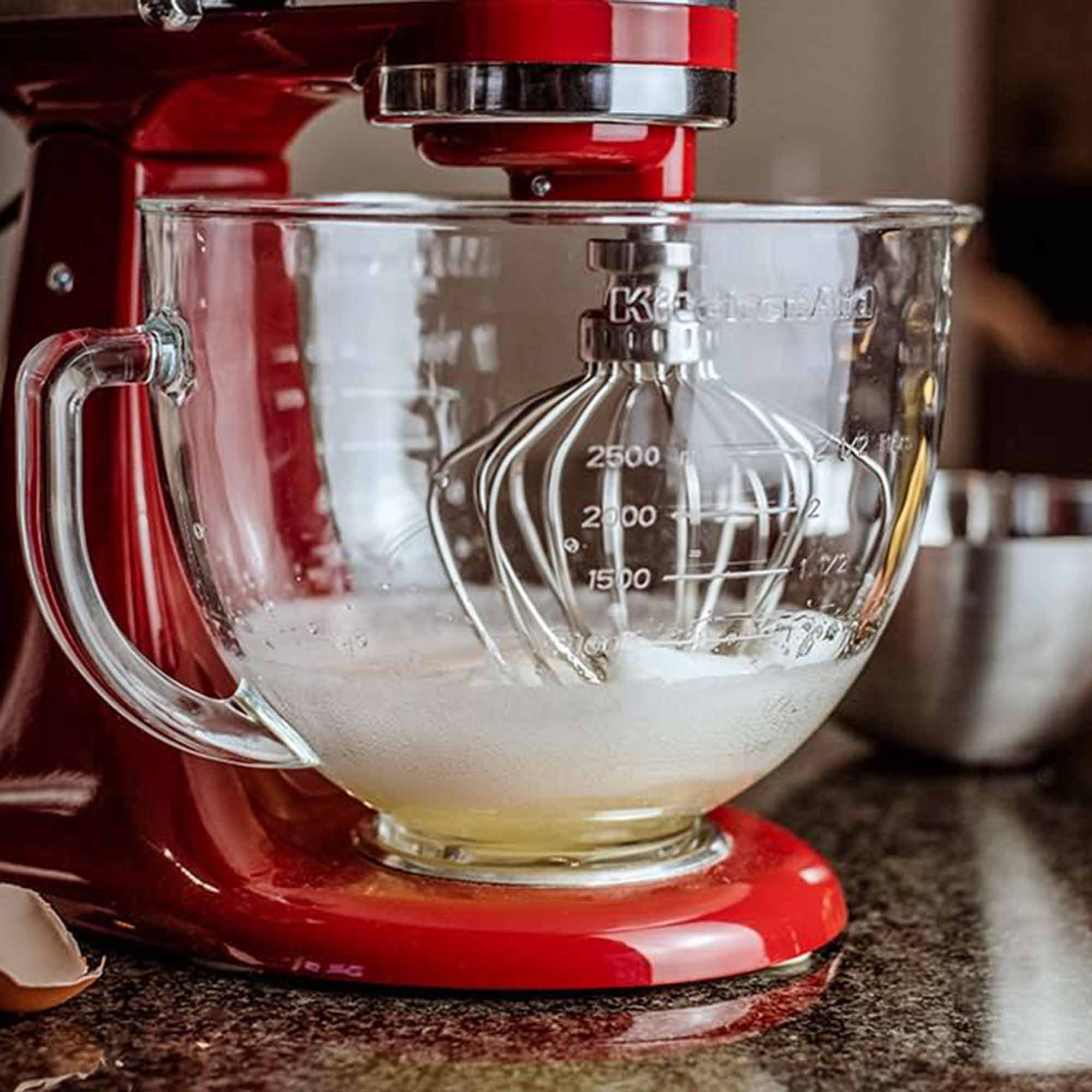 Ein roter KitchenAid-Mixer mit Glasschüssel und Schneebesenaufsatz. Die Schüssel enthält Eiweiß.