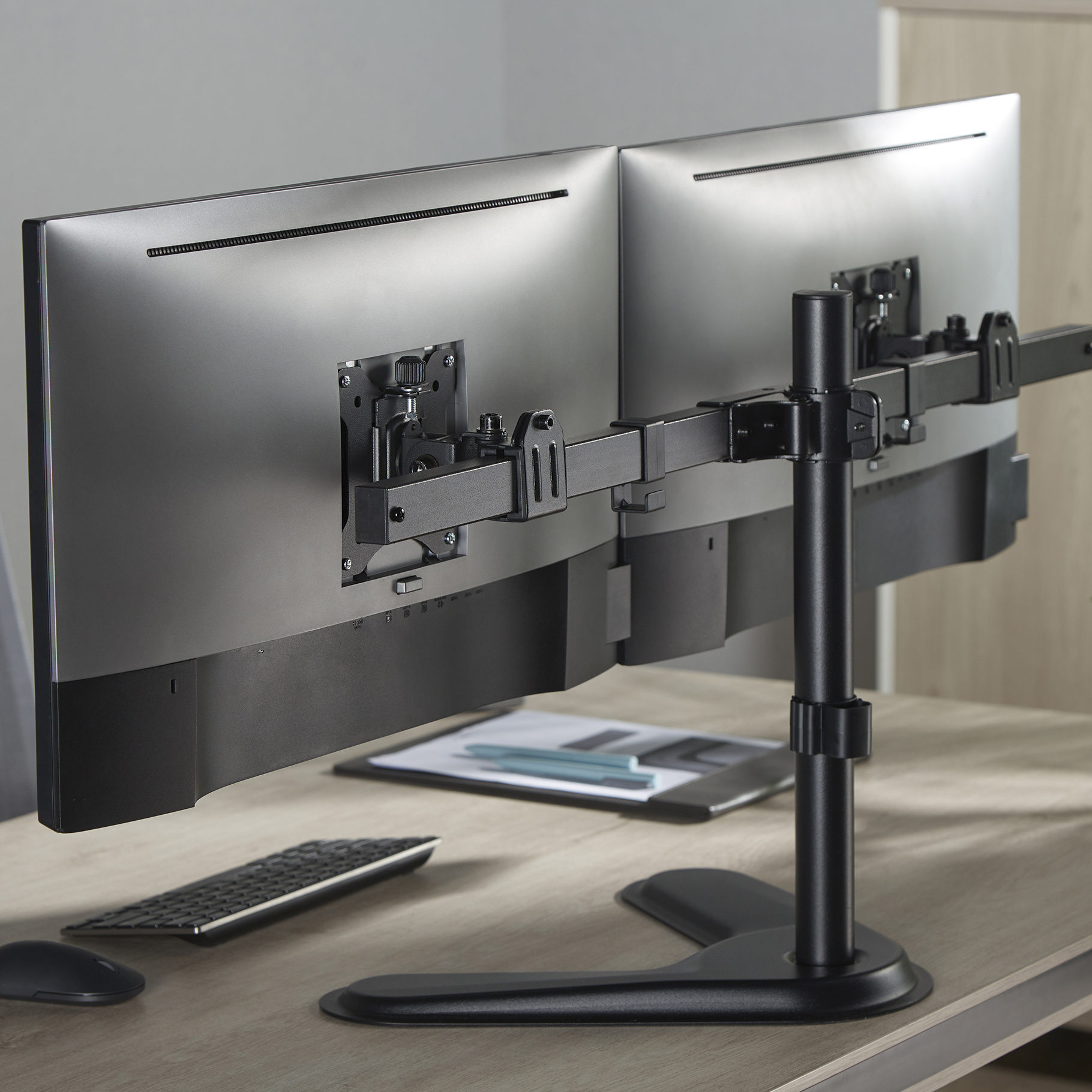 Dubbele monitoren op standaard, toetsenbord en muis op houten bureau, met klembord op de achtergrond.