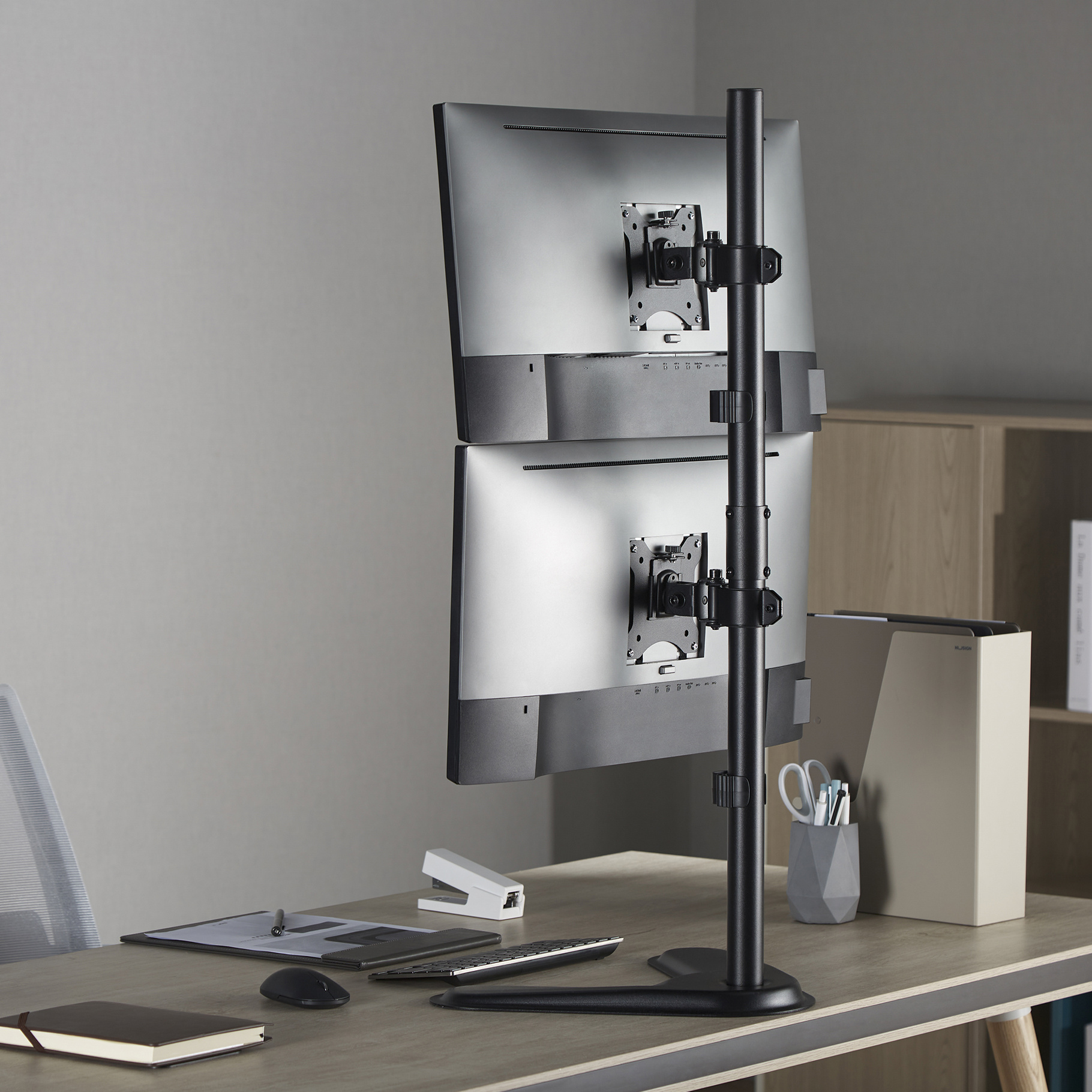 Twee computermonitoren op een zwarte standaard op een bureau. Bureau heeft een nietmachine.