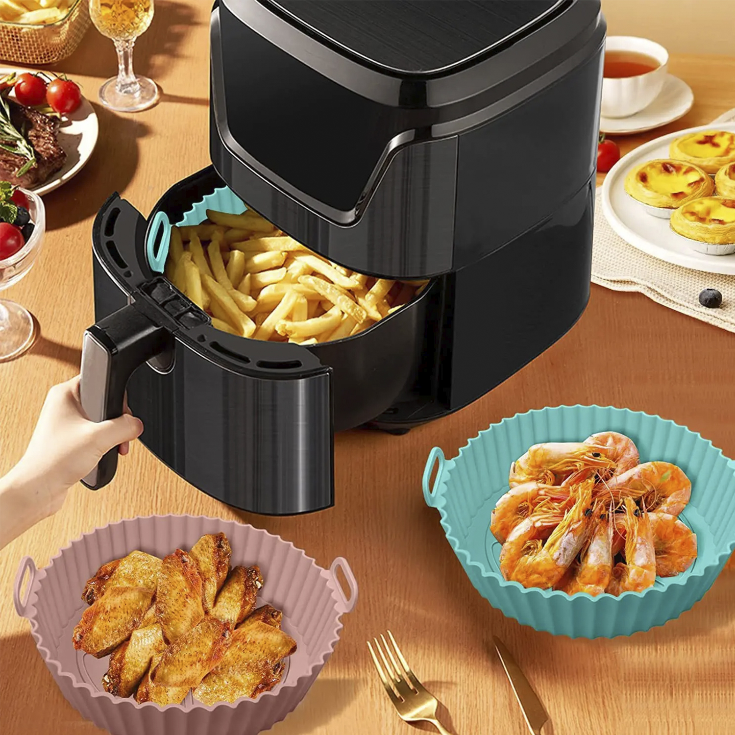 Una friggitrice ad aria con cibi fritti e due vassoi in silicone con cibo su un tavolo di legno.
