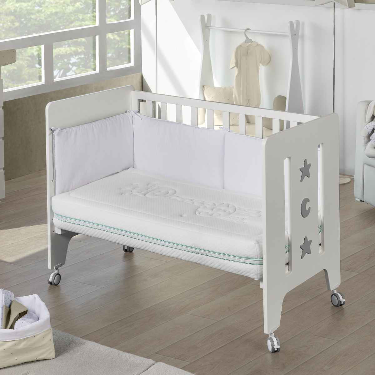 Una cuna blanca con colchón, ropa de cama blanca y protector. Suelo de madera clara.