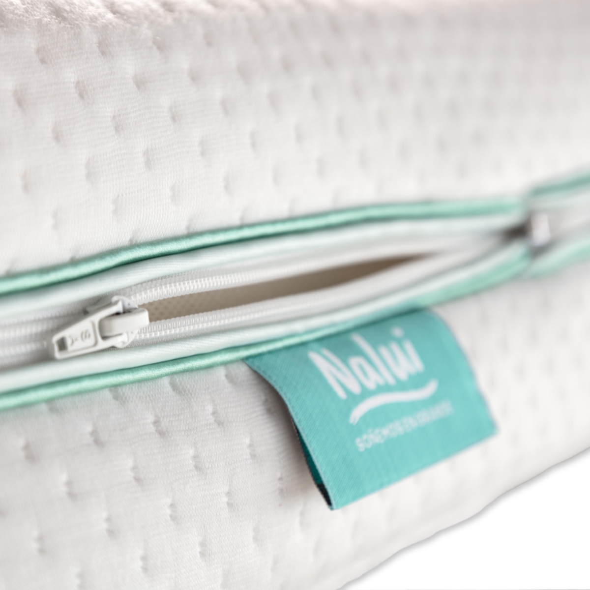 Primer plano de una almohada blanca con cremallera. Una etiqueta turquesa dice 'Nalu'. La cremallera está abierta.