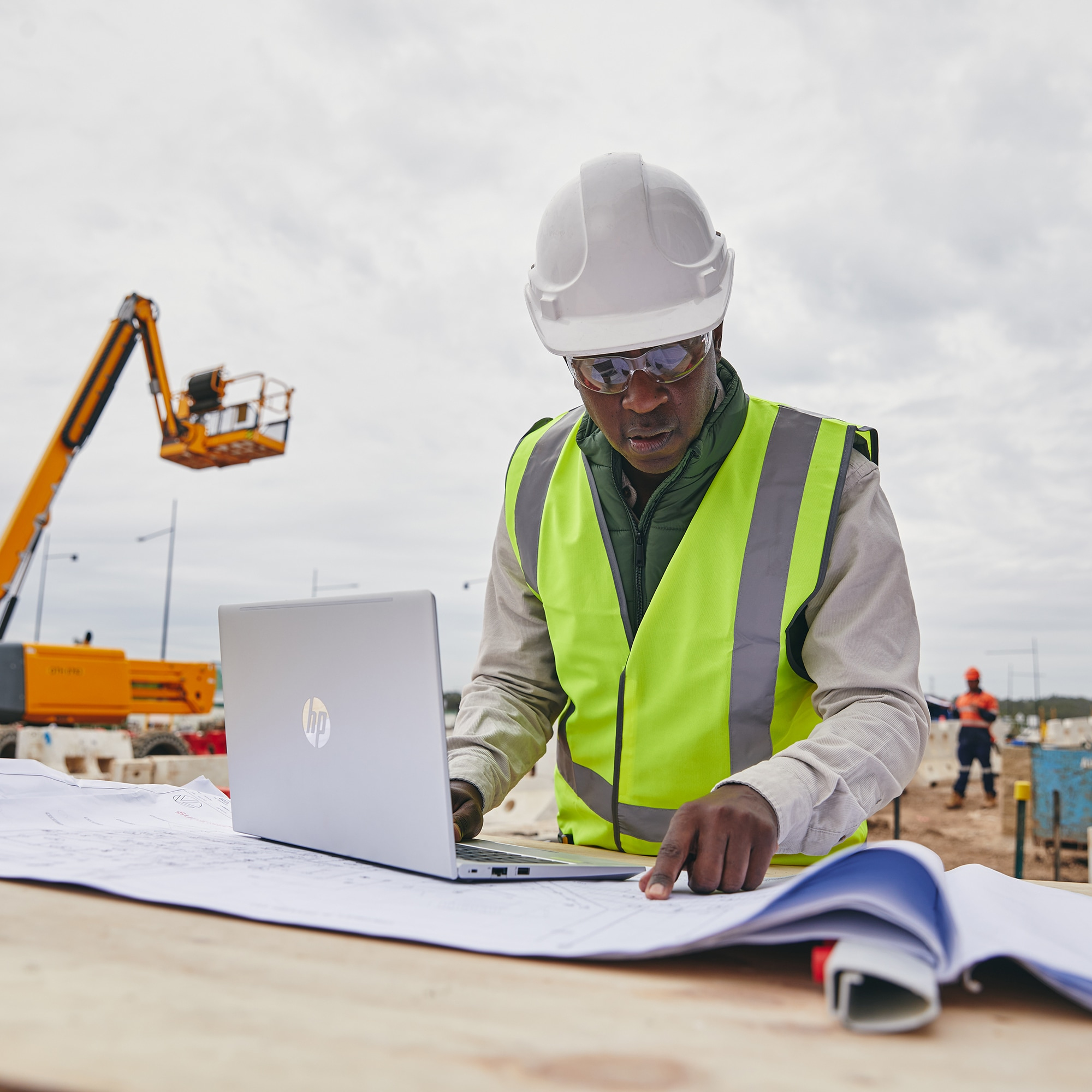 Uomo con gilet giallo e casco che lavora su un laptop in un cantiere. Progetti visibili.