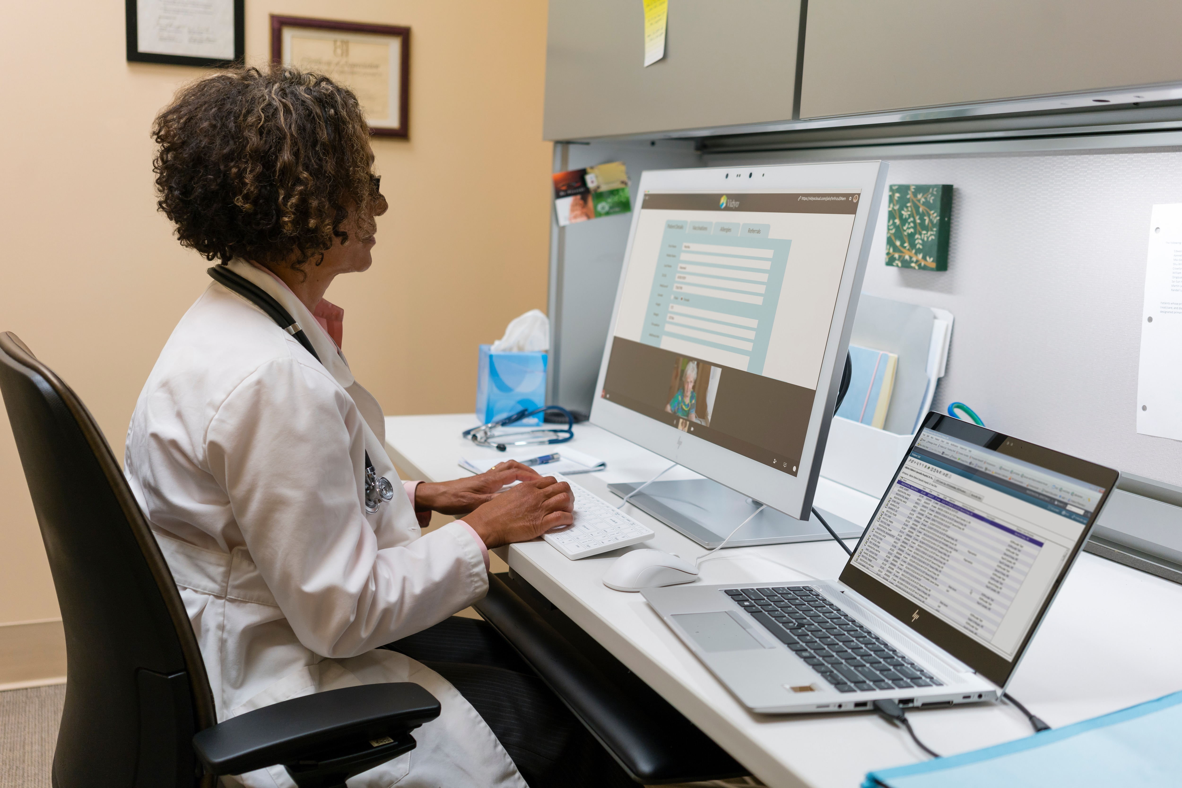 Il medico è seduto alla scrivania a digitare. Due monitor mostrano un modulo paziente. Anche un laptop è aperto.