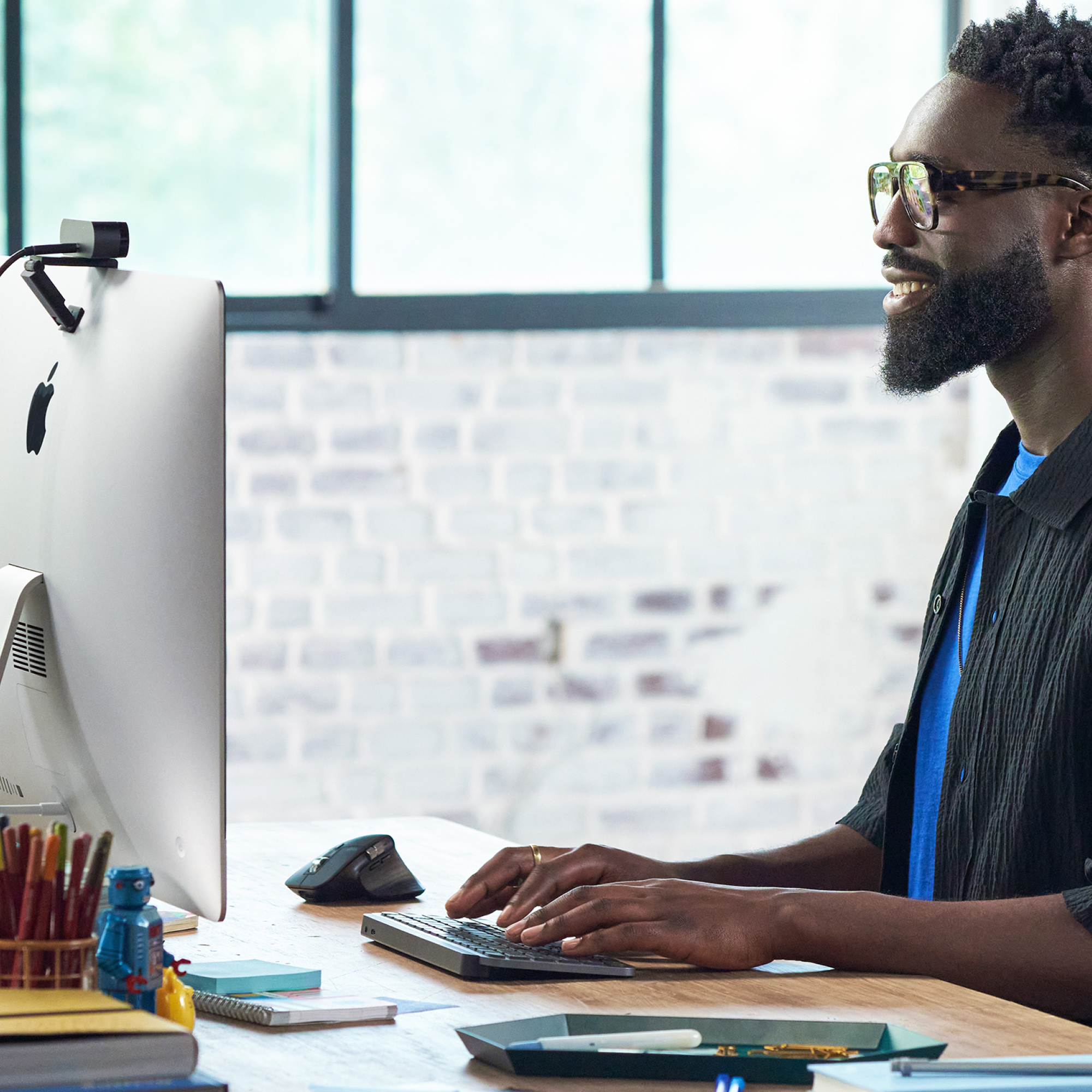 A man with glasses smiles while typing on a keyboard, with a computer screen and desk items.