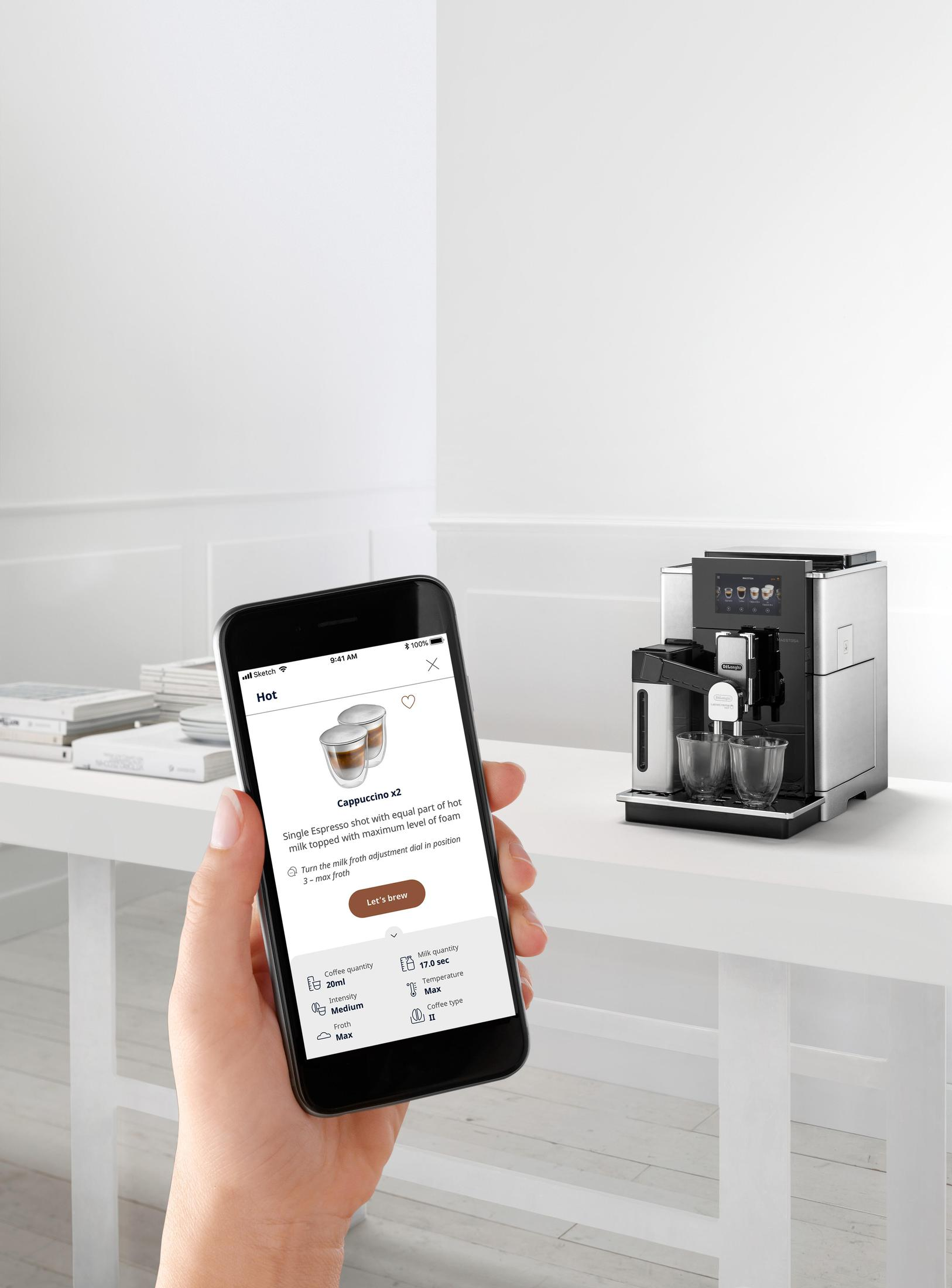 Die Hand einer Person hält ein Telefon mit einer Kaffeemaschinen-App; eine Kaffeemaschine steht auf einem weißen Tisch.