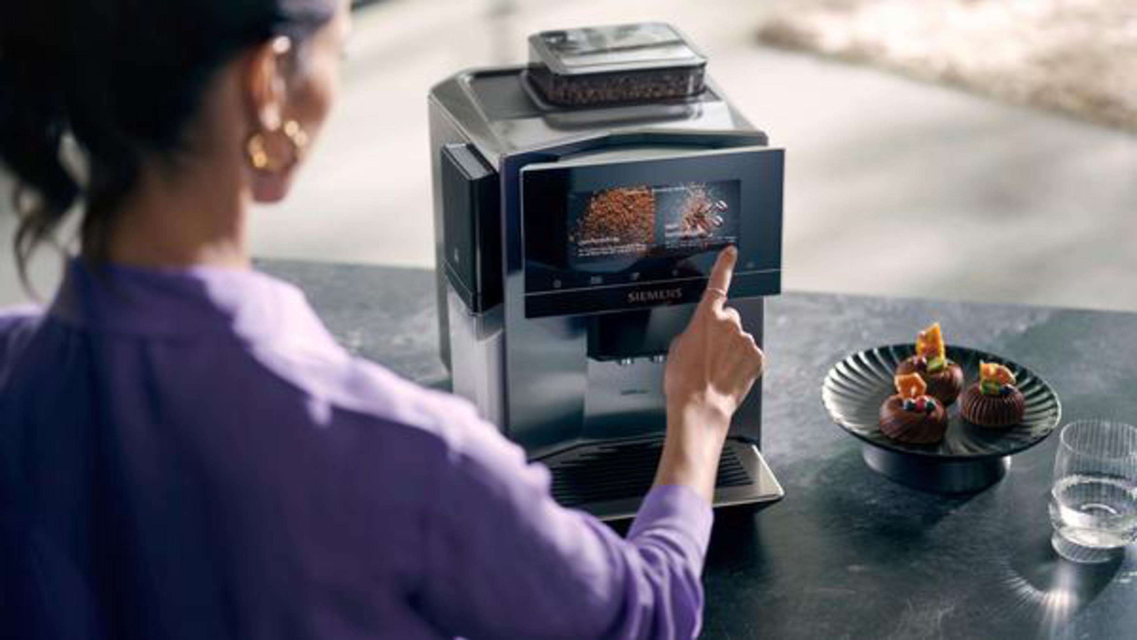 Kobieta używa ekspresu do kawy. Maszyna ma ekran, którego dotyka. Talerz z ciastkami.