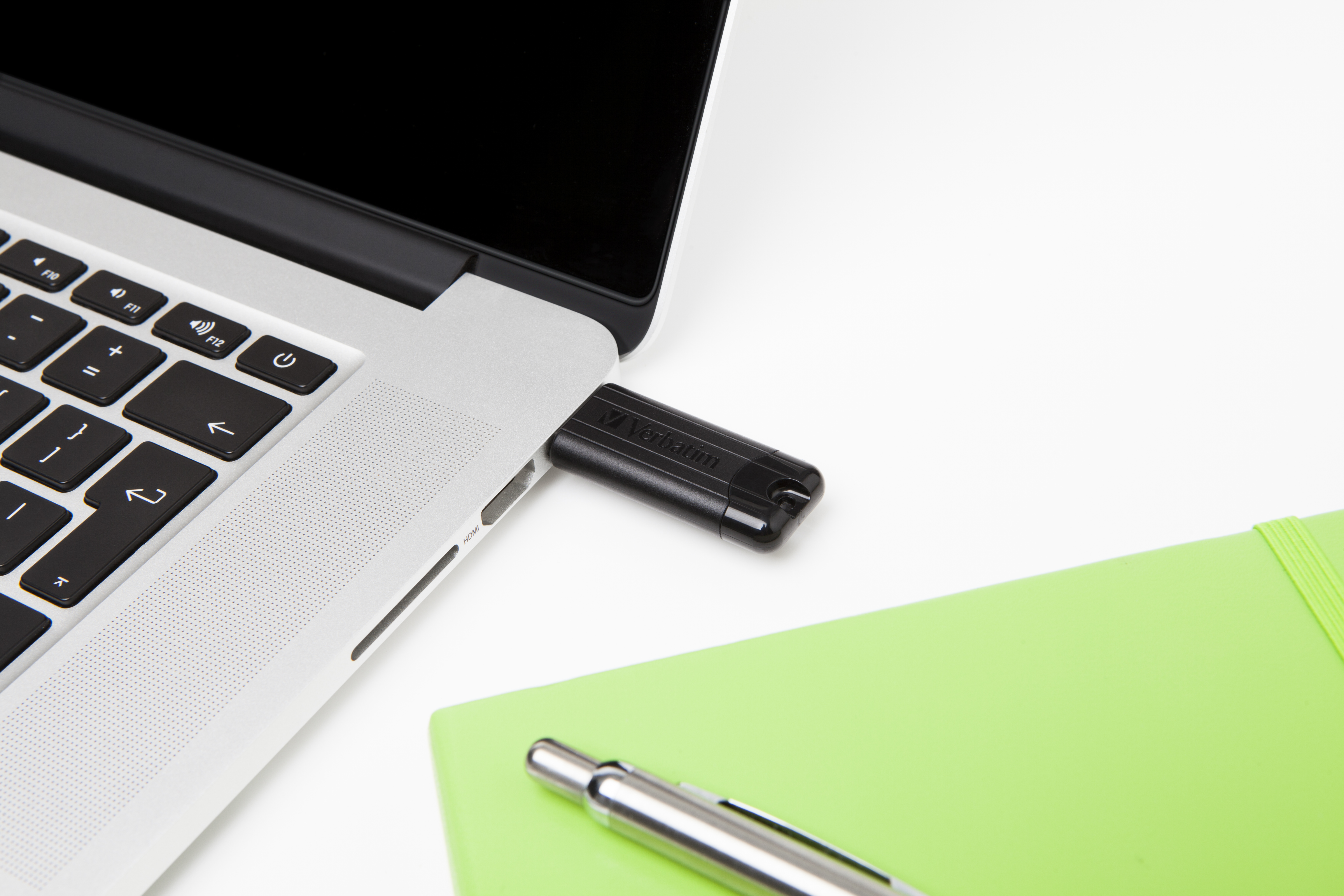 Laptop z włożonym czarnym pendrive'em, obok zielonego notatnika i długopisu.