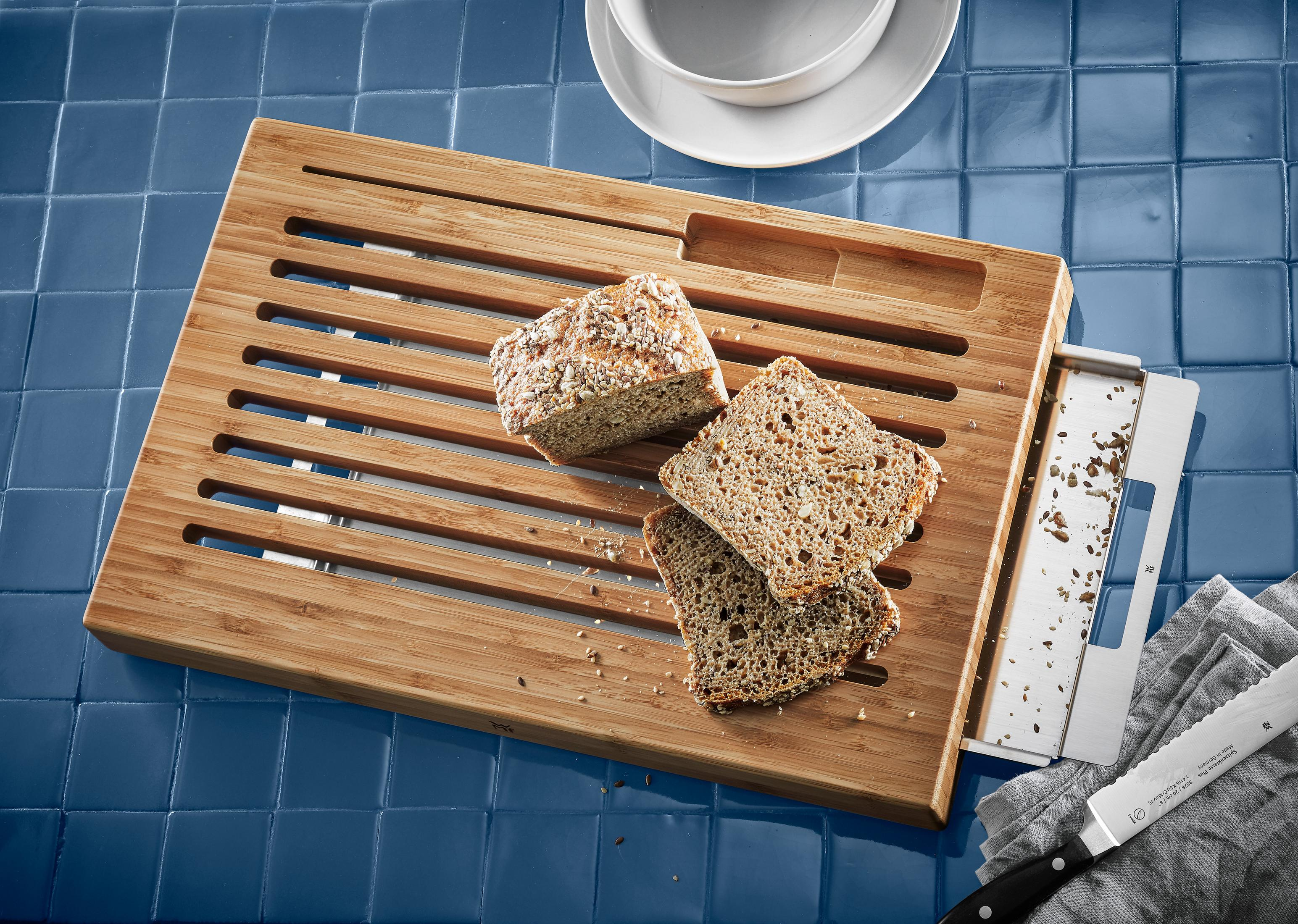 Holz-Schneidebrett mit geschnittenem Brot, Krümeln und einem Messer auf blauer Fliesenoberfläche.