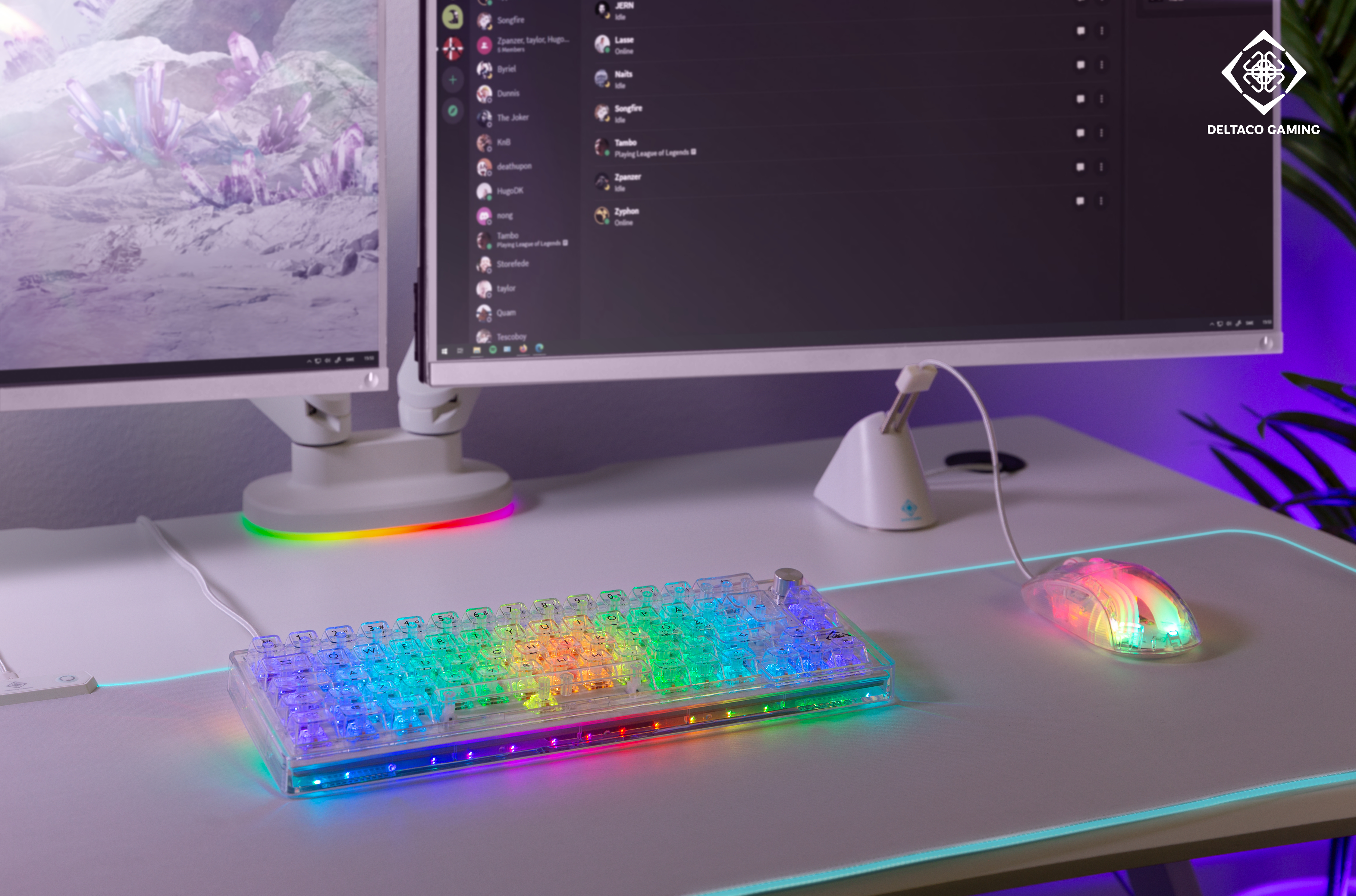 Configurazione di gioco: tastiera e mouse illuminati, doppi monitor con Discord, scrivania bianca, dettagli viola.