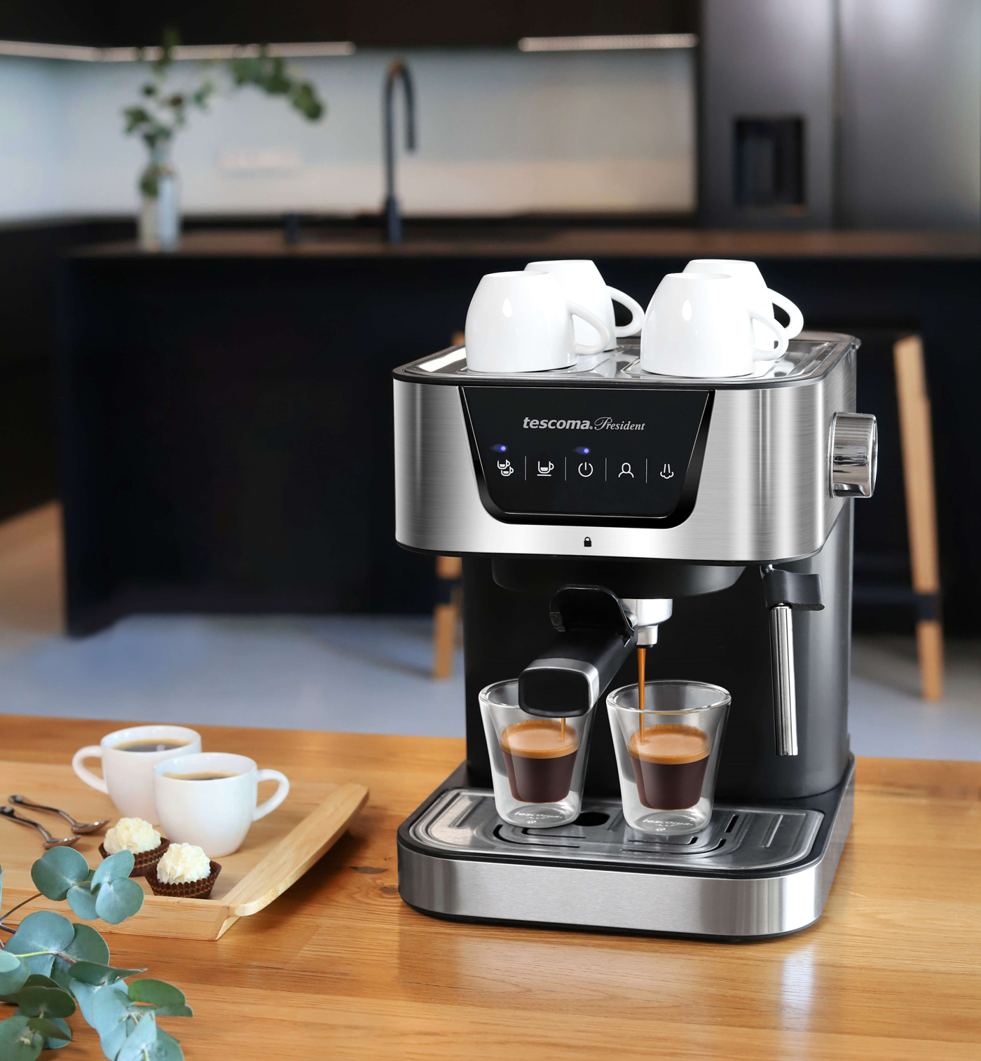 Espressomaschine mit zwei Kaffeetassen, drei Tassen oben. Holztisch mit Gebäck und grünen Zweigen.