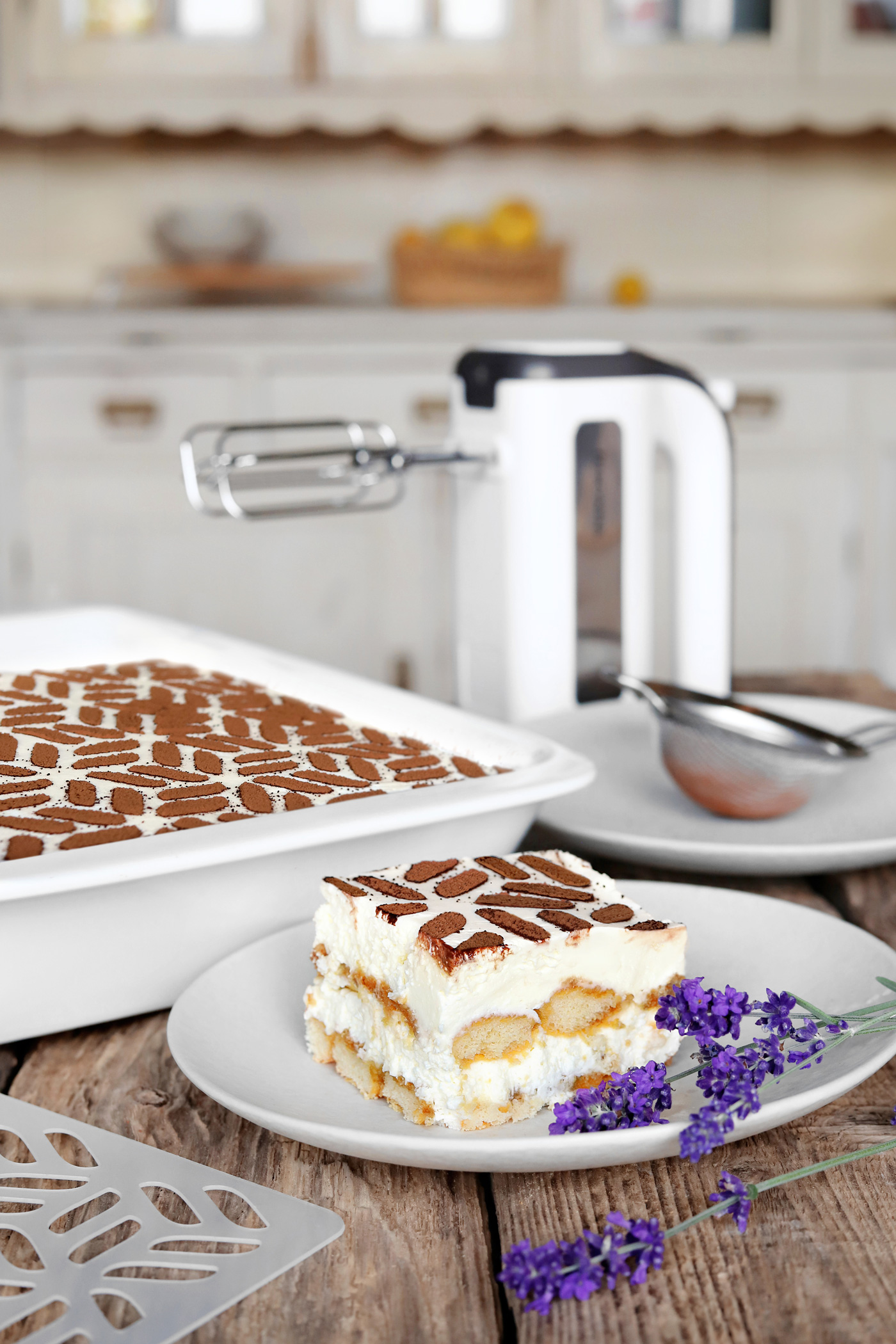 Tiramisu auf einem Teller, Kakaopulver und ein Handmixer. Rustikaler Holztisch und Küche im Hintergrund.