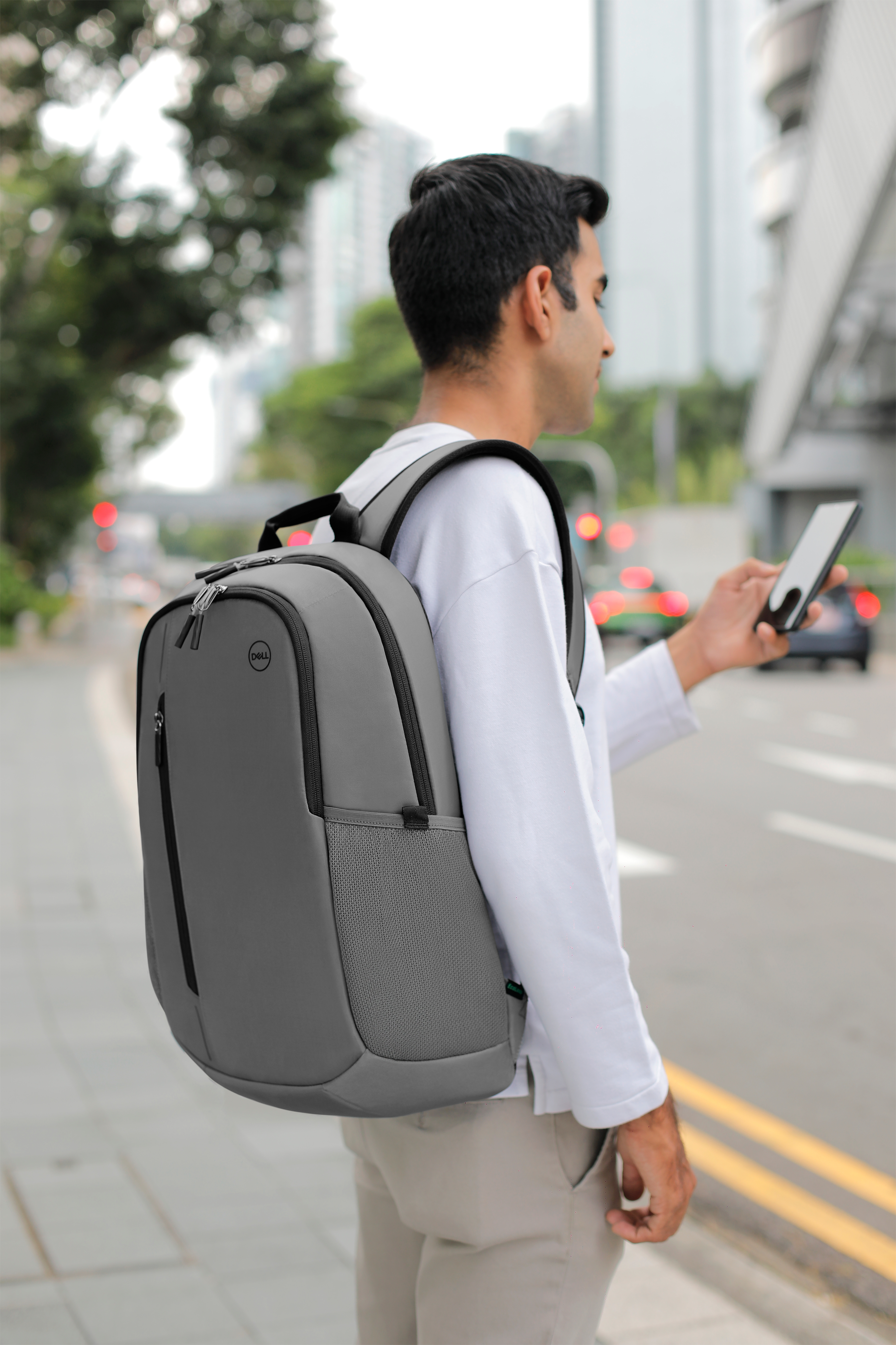 Man wearing a grey backpack and holding a phone, walking on a city street.