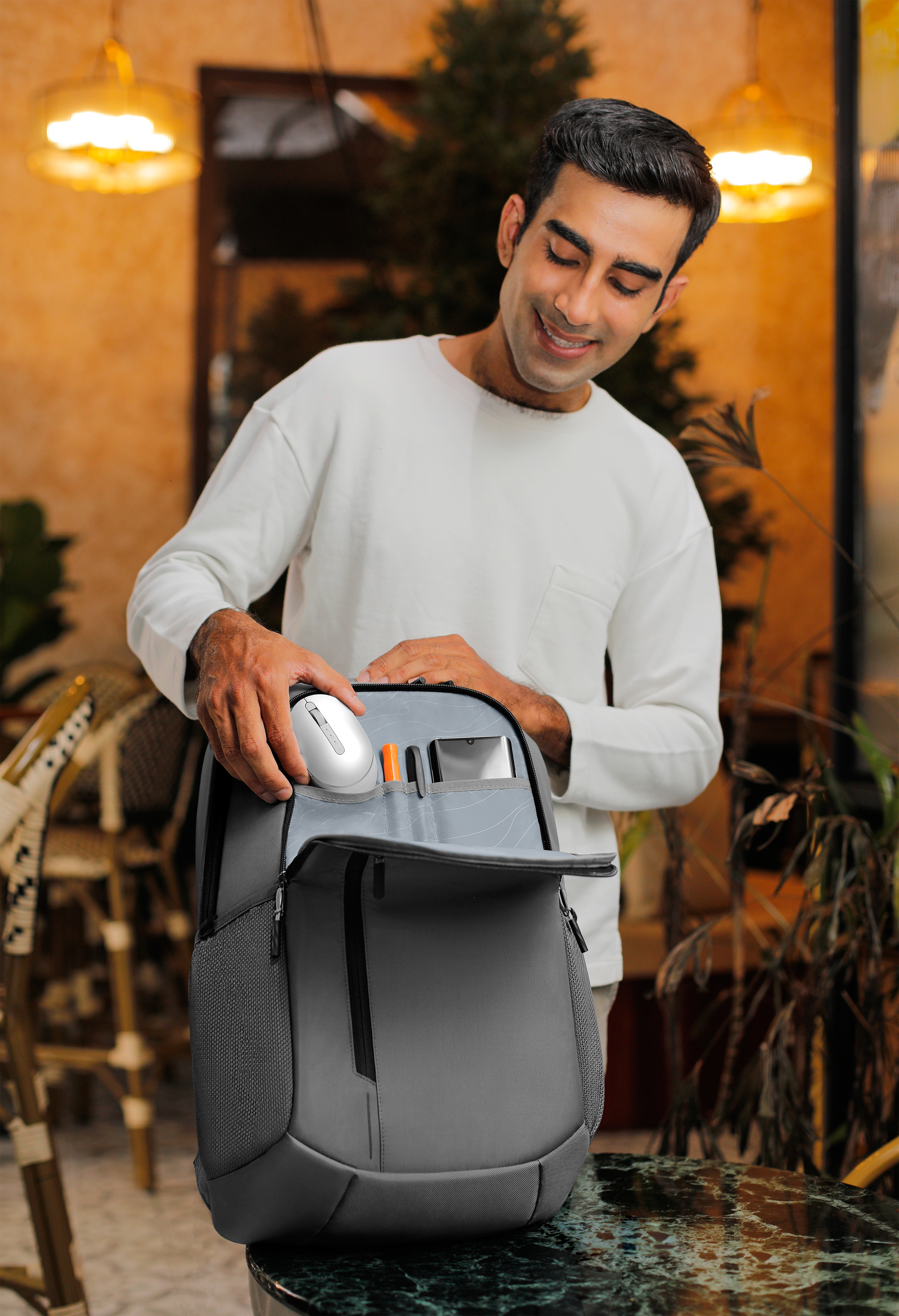 Man opening a gray backpack. Inside are a mouse, pens, and tablet. Background is a cafe setting.