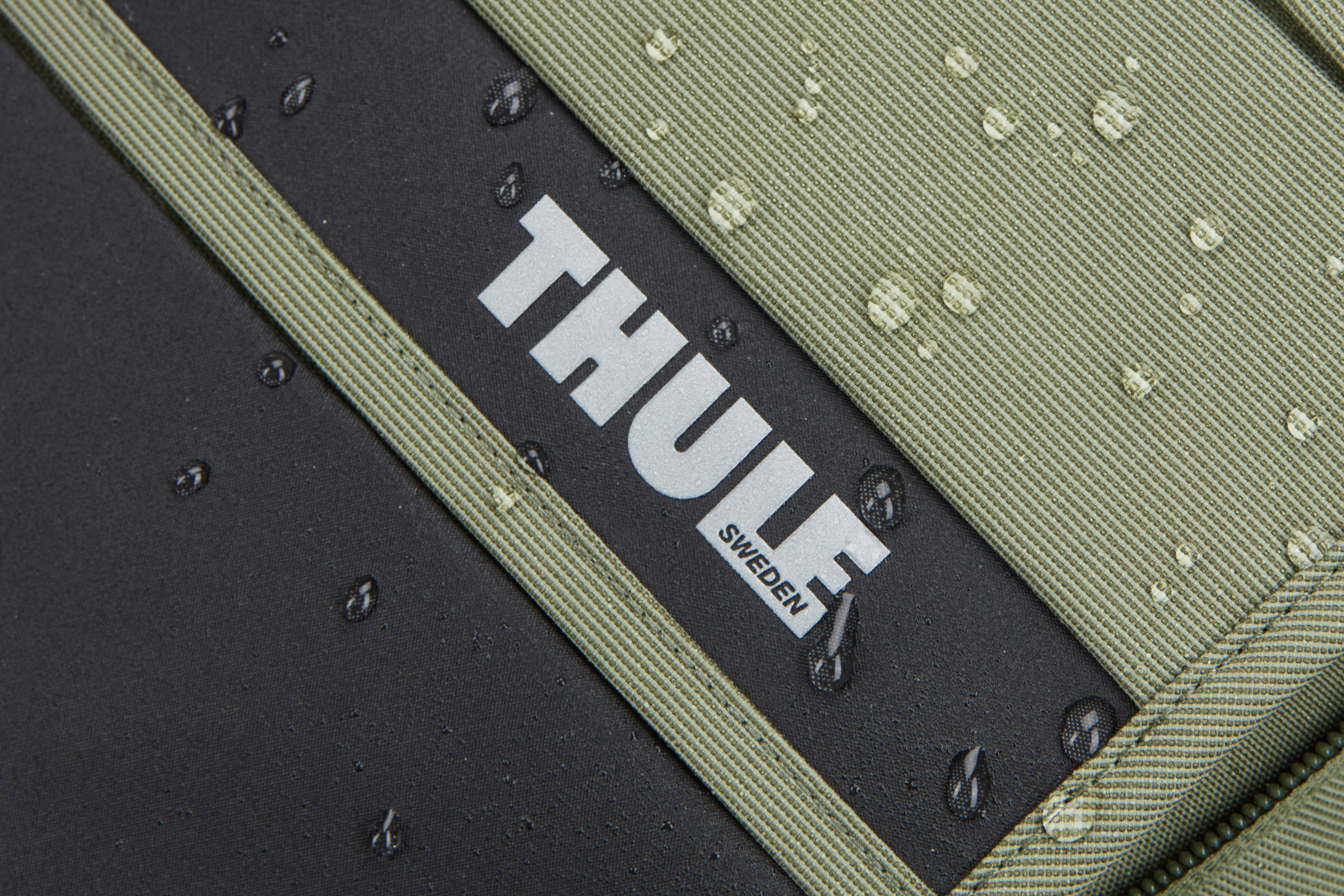 Nahaufnahme einer schwarzen und olivgrünen Tasche mit Thule-Logo und Wassertropfen.