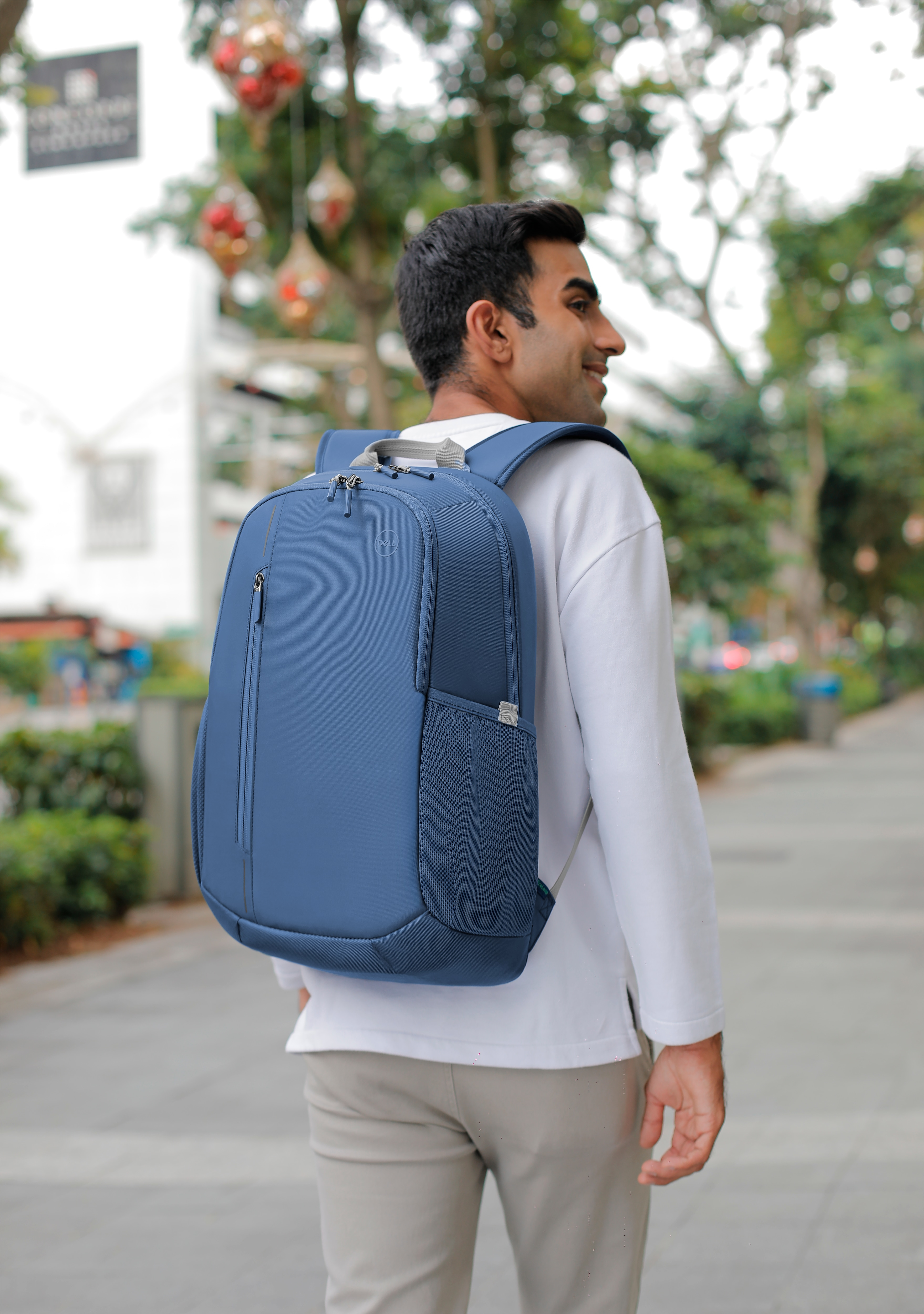 Mann mit blauem Rucksack, der auf einem Bürgersteig geht. Bäume und Gebäude im Hintergrund.