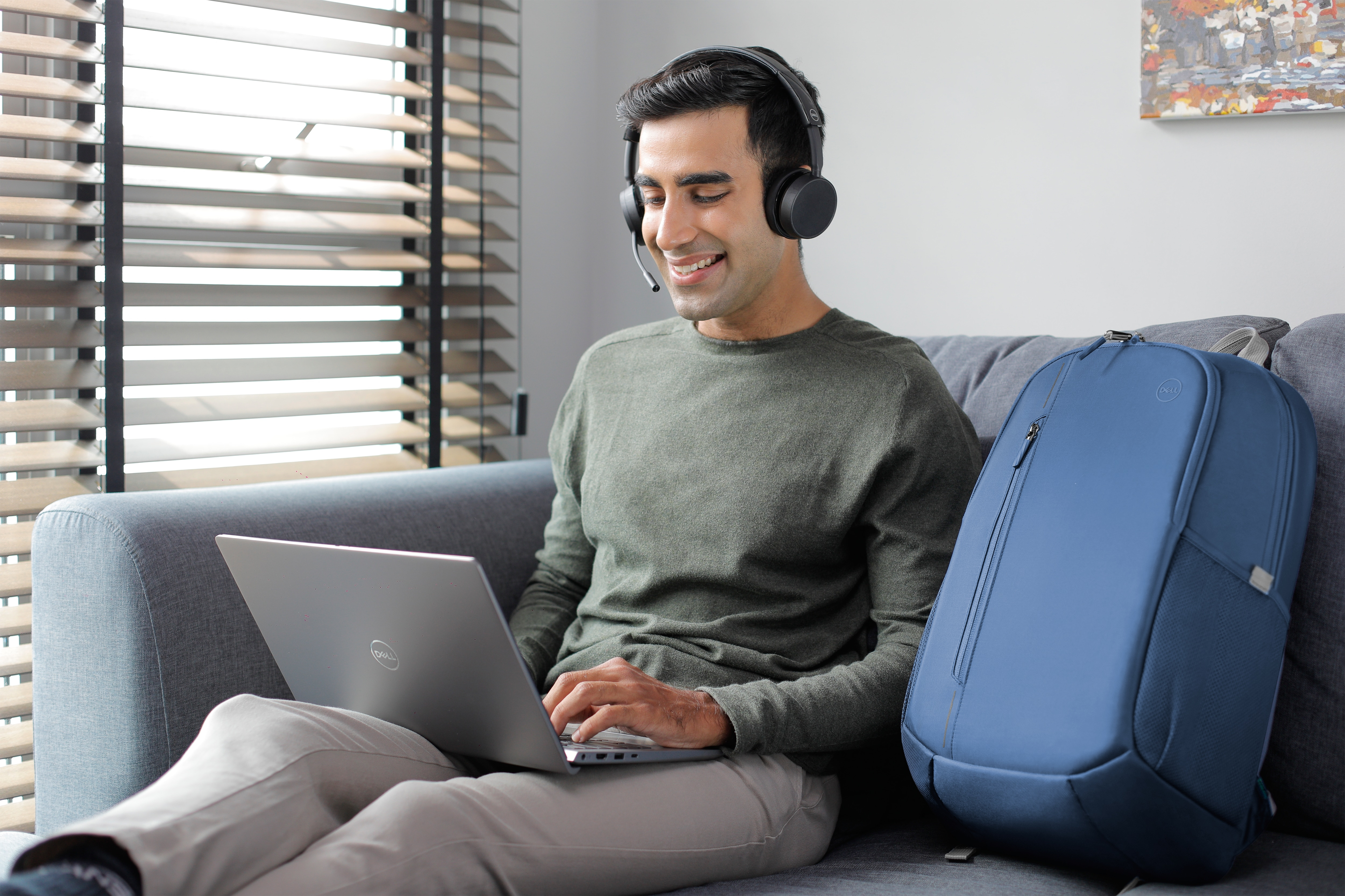 Mann auf einem Sofa mit Laptop, trägt Headset, lächelt. Blauer Rucksack daneben.